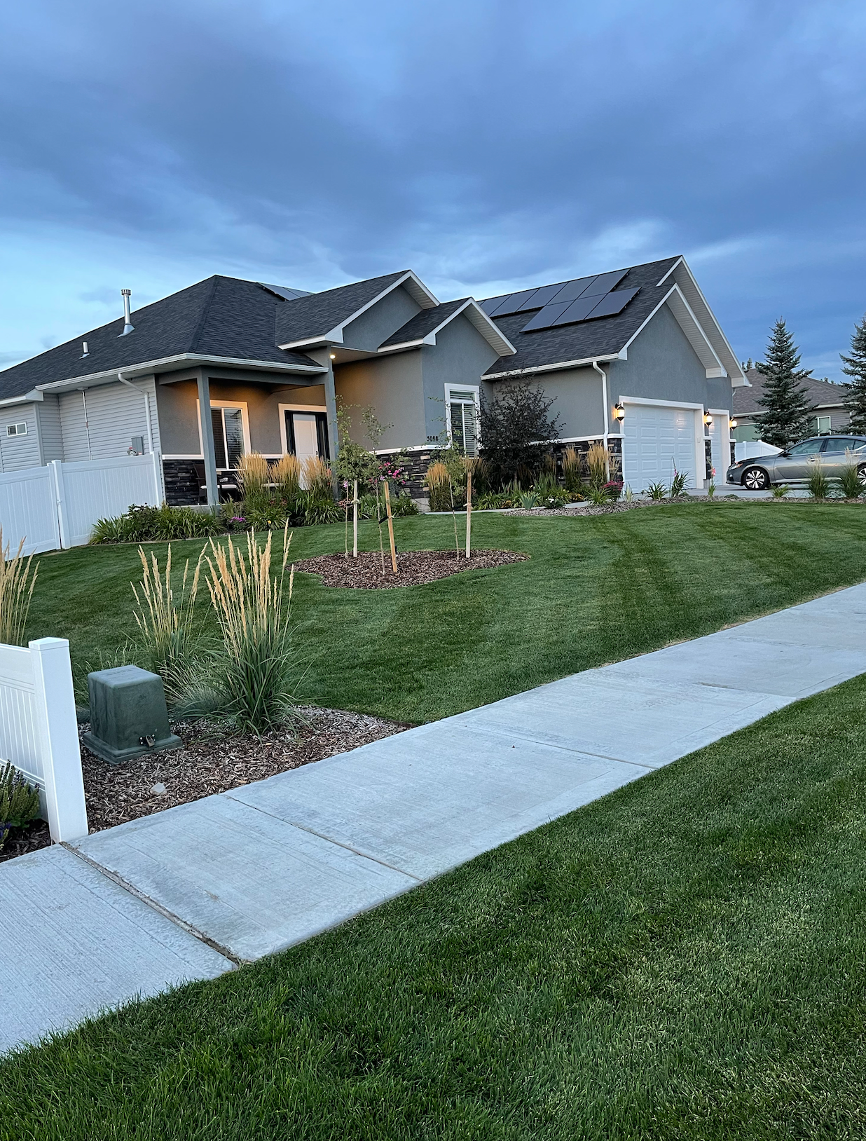A gray, single-story suburban house with a dark roof and solar panels, viewed from the sidewalk on a cloudy day.