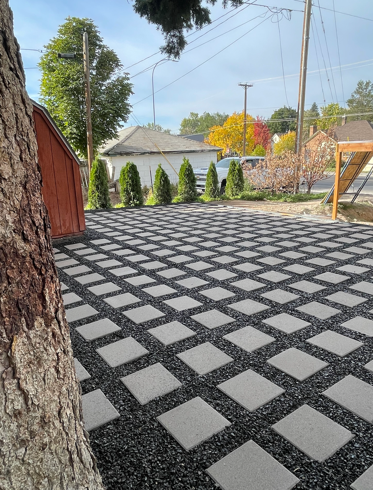 A grid of square concrete pavers set in black gravel, next to a large tree and a small garden with a red shed.