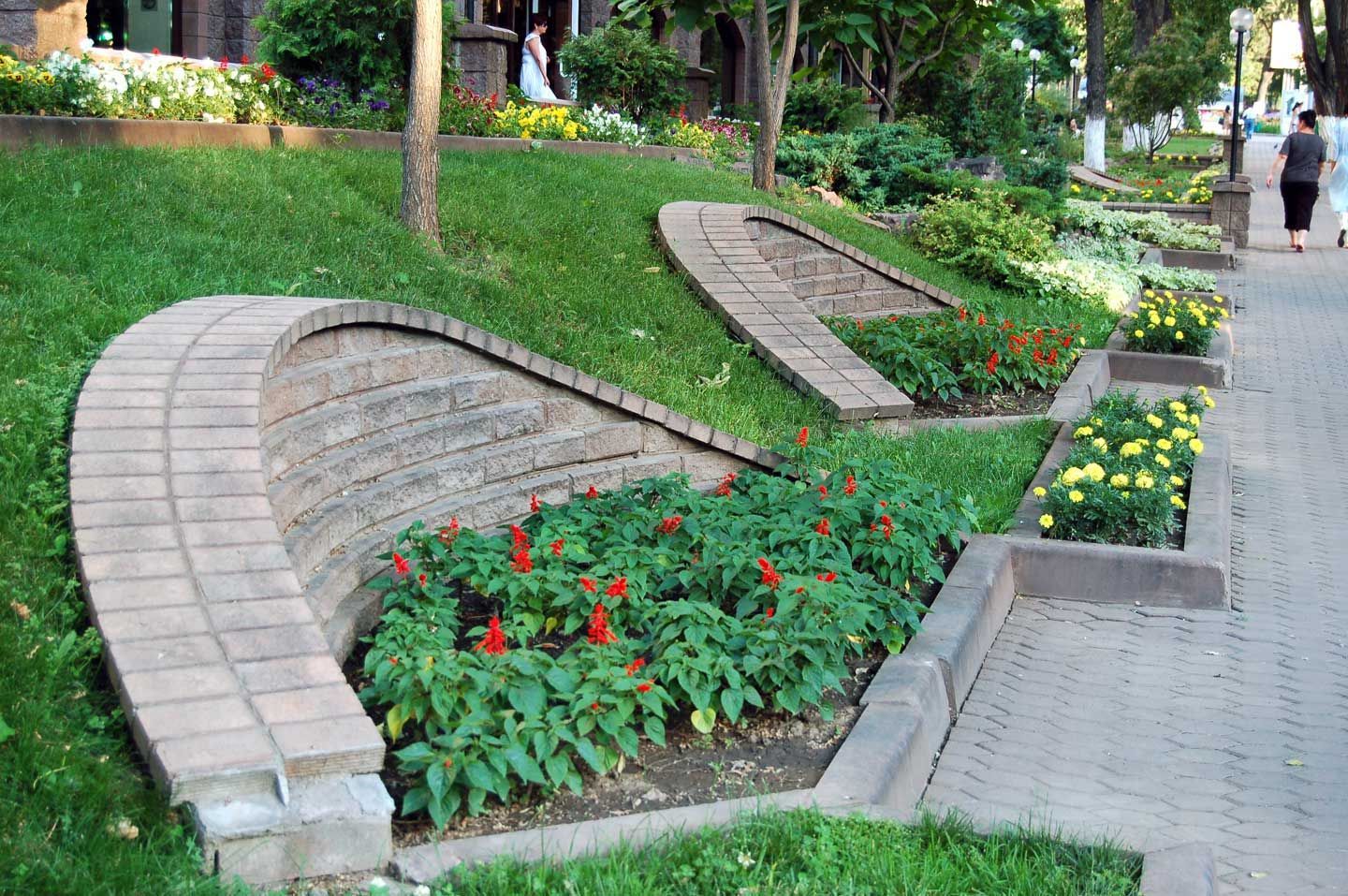 Brick-edged flower beds on a sloping green lawn, red and yellow flowers, person walking on a paved path.