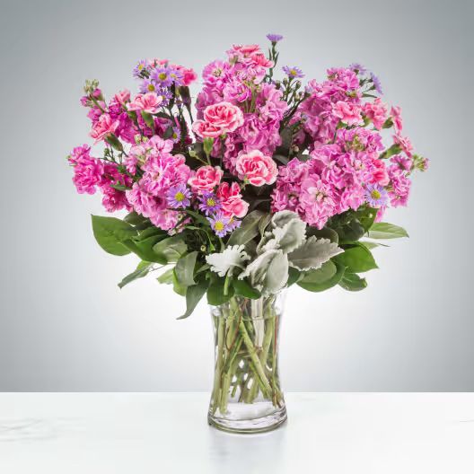 A clear glass vase filled with a vibrant arrangement of pink carnations, purple statice, and silver-green dusty miller.