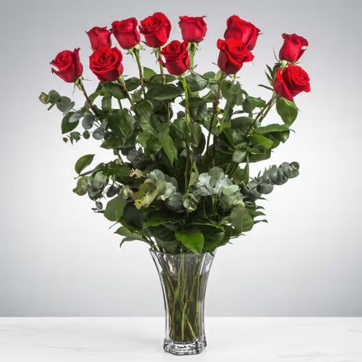 A bouquet of red roses with green eucalyptus leaves in a clear glass vase against a plain, light background.