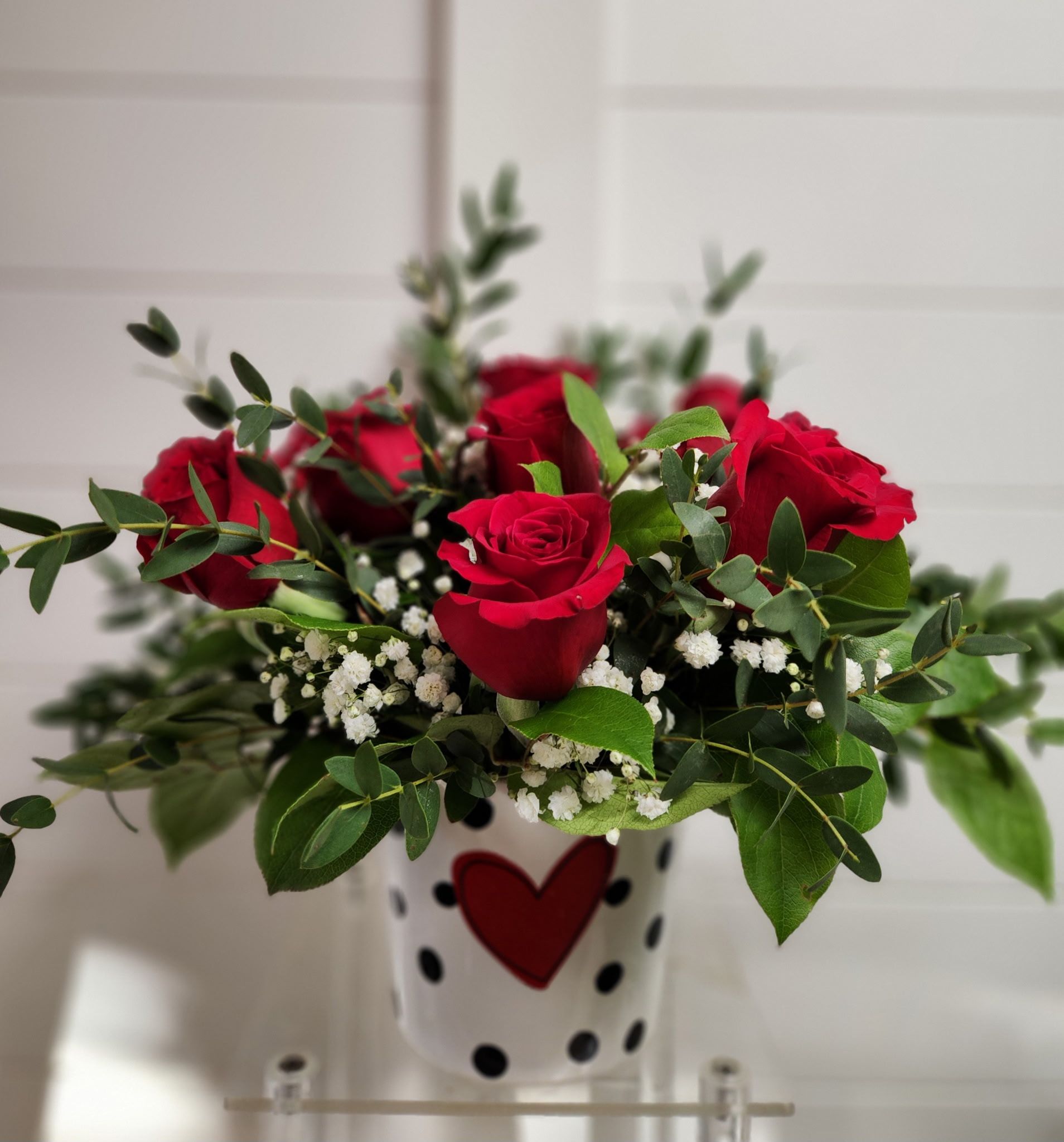 A bouquet of red roses, greenery, and baby's breath in a white planter decorated with a red heart and black polka dots.