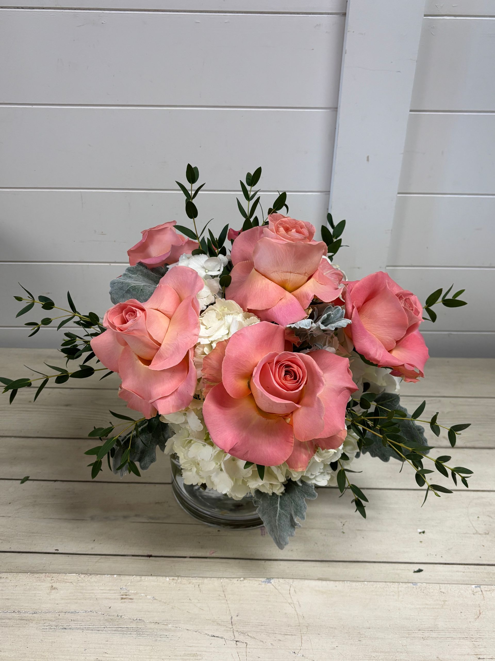 A glass vase filled with large, peach-colored roses, white hydrangea, and greenery against a white wooden wall.