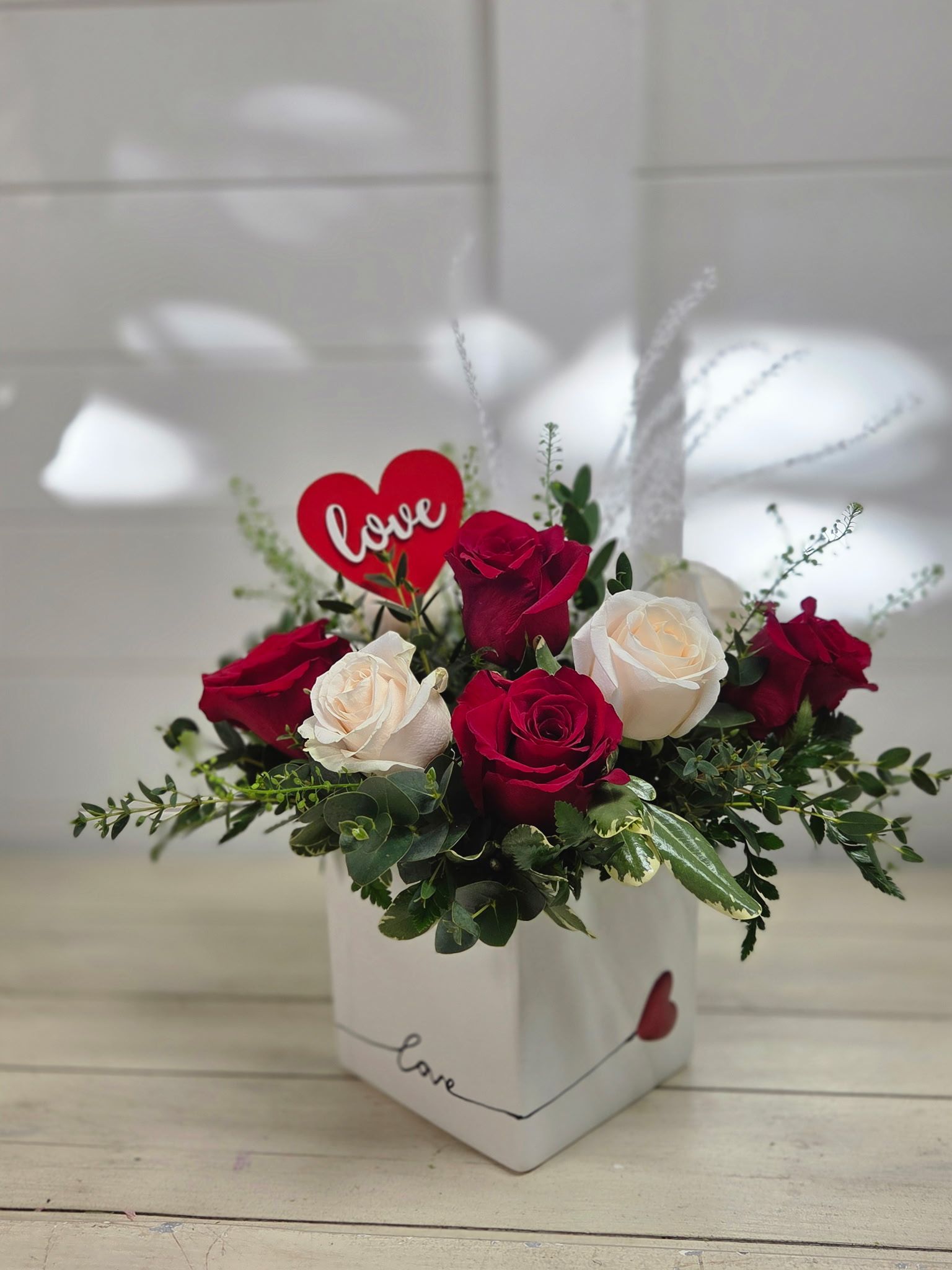 A square white vase decorated with a red heart holds a bouquet of red and white roses with greenery and a 