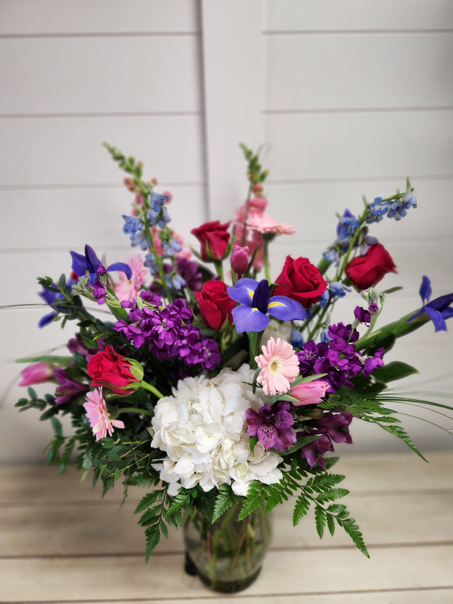 A vibrant floral arrangement in a glass vase featuring white hydrangeas, red roses, purple irises, and blue larkspur.