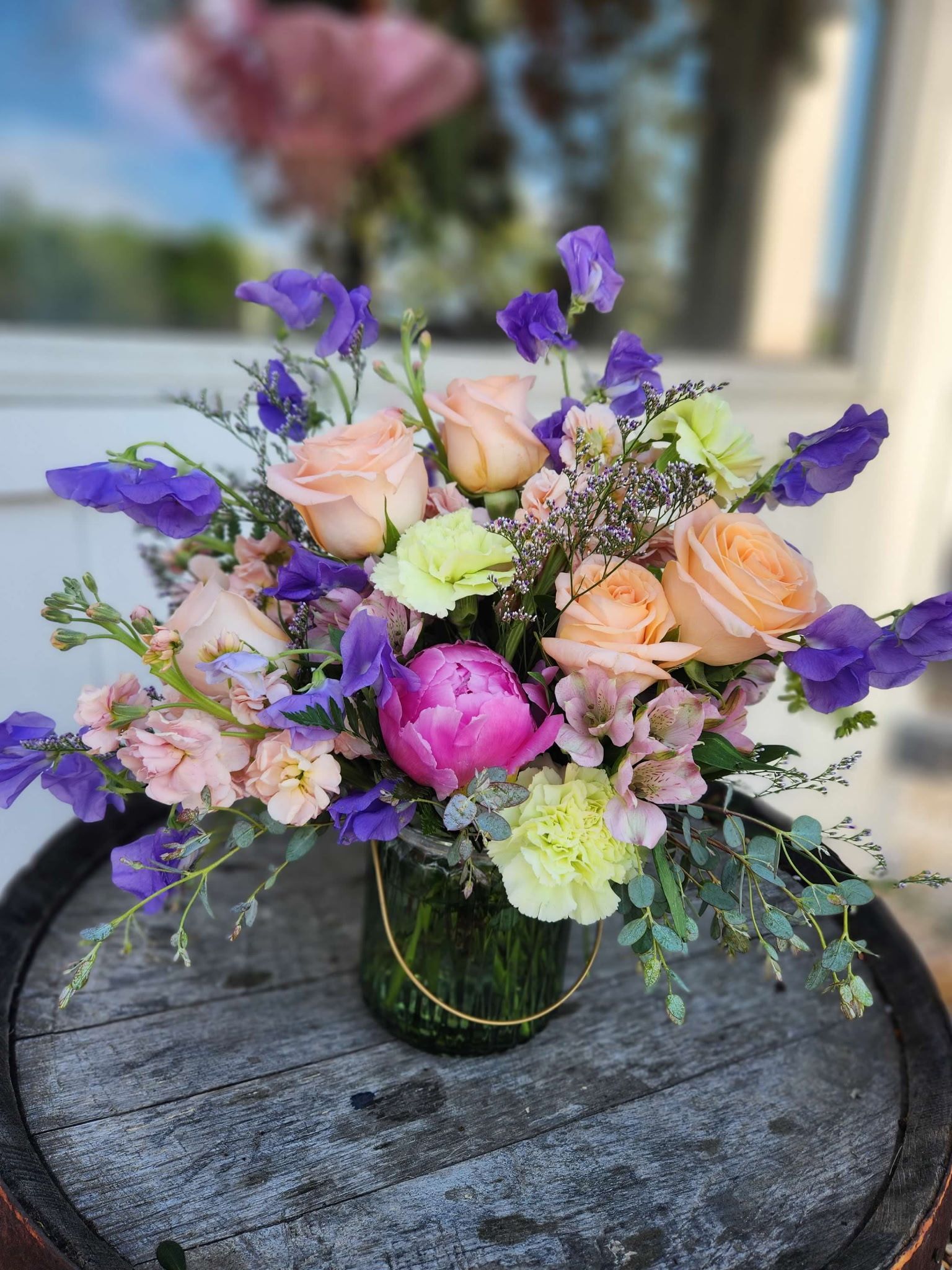 A glass vase of flowers including peach roses, a bright pink peony, and purple sweet peas, resting on a wooden barrel.