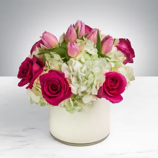 A bouquet of pink roses and tulips mixed with green hydrangea in a simple white cylindrical vase against a gray background.