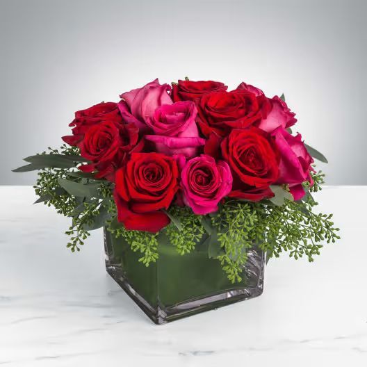 A square glass vase filled with a lush arrangement of red and bright pink roses accented with greenery on a white surface.
