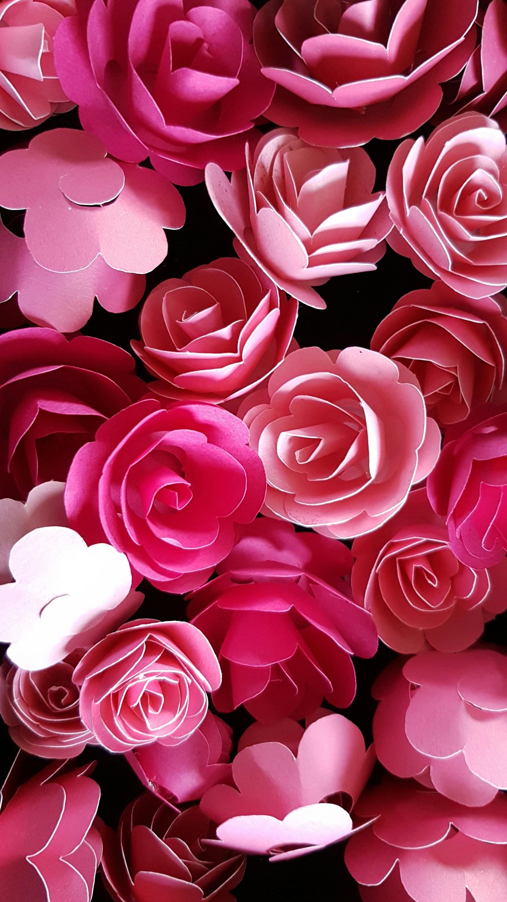 A dense arrangement of paper roses in shades of pink, ranging from light pastel to deep magenta, against a black background.