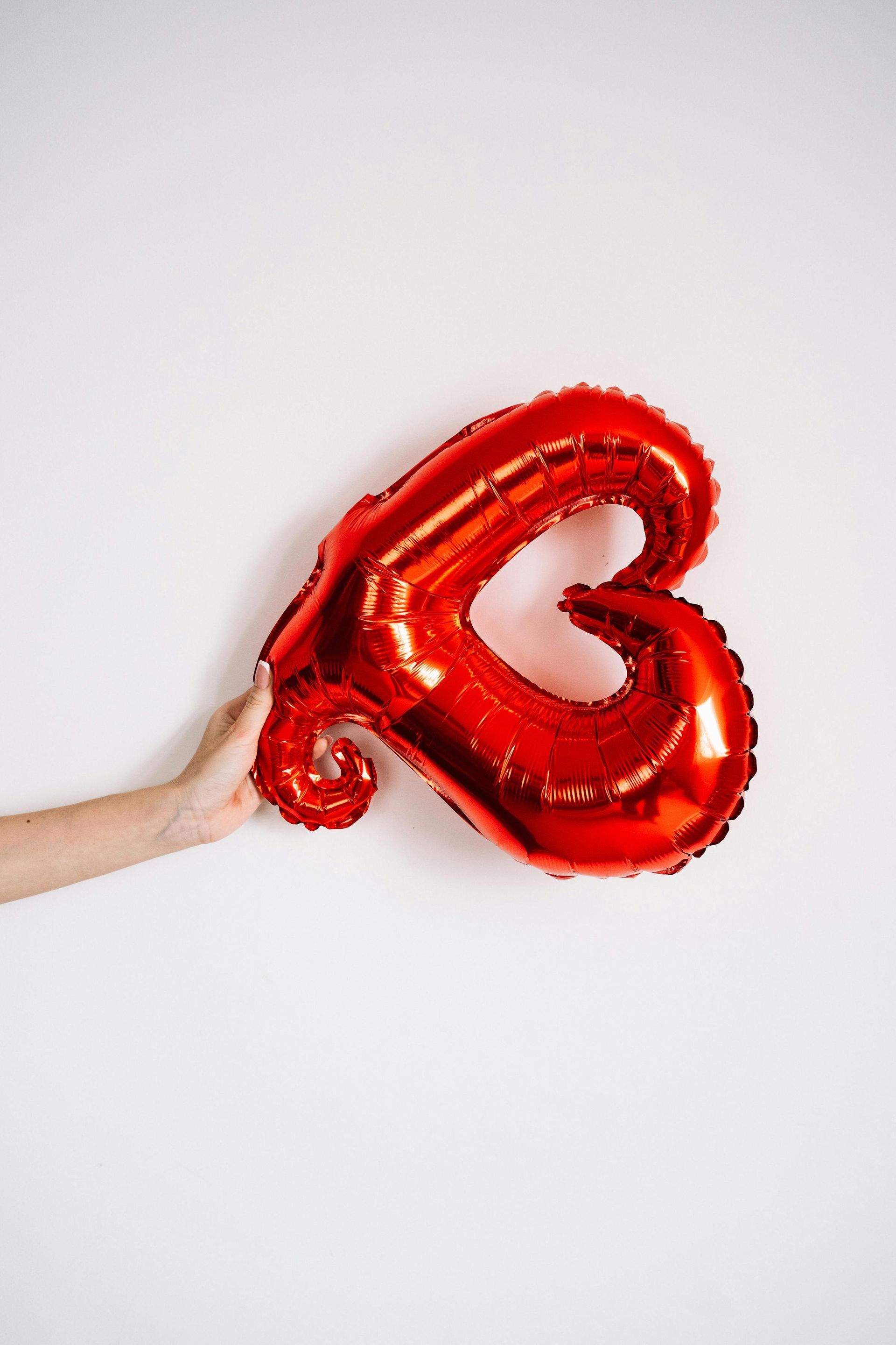A person’s hand holds a shiny, red, heart-shaped balloon against a plain white background.