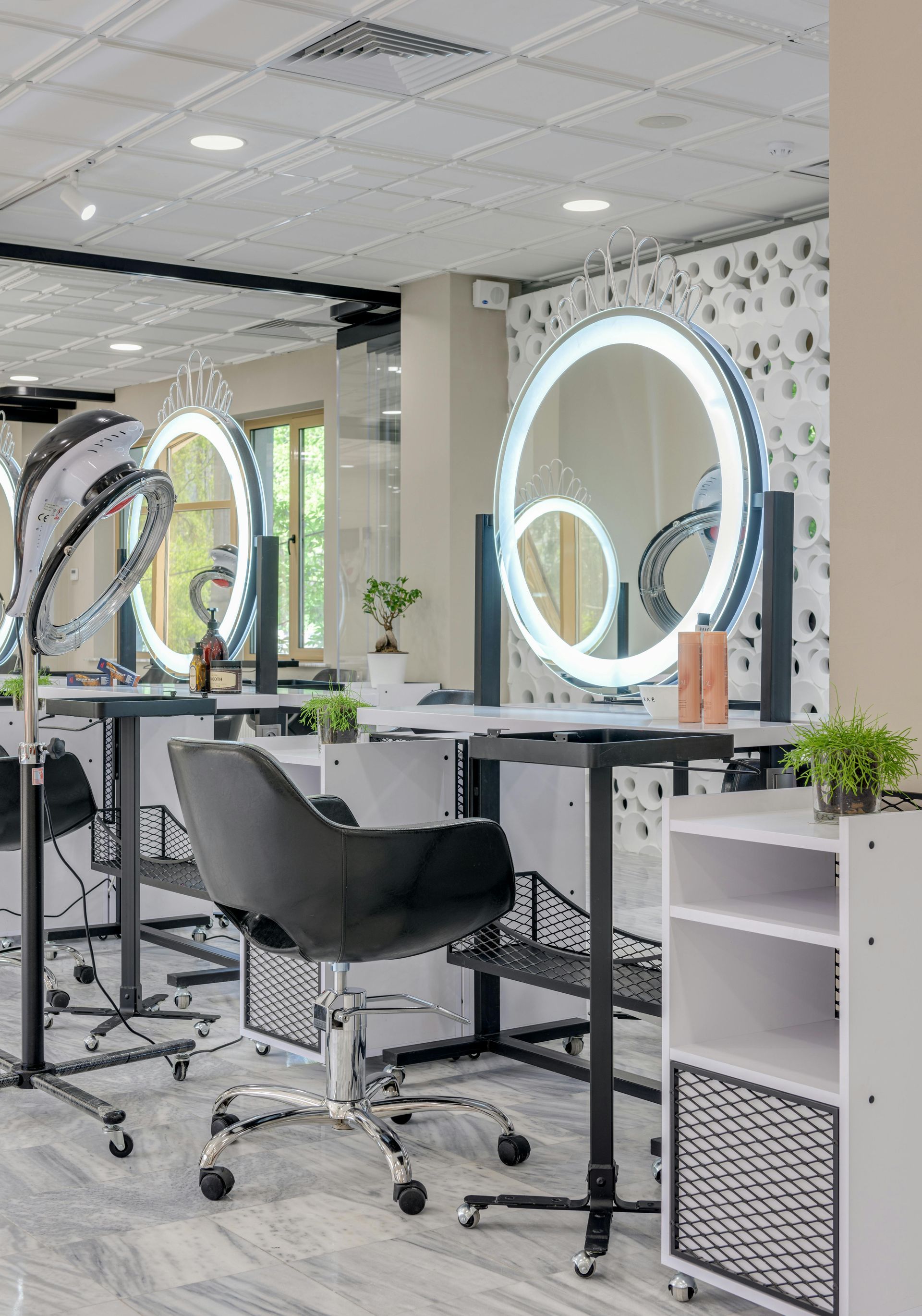 A modern hair salon featuring black styling chairs, circular illuminated vanity mirrors, and white storage cabinets.