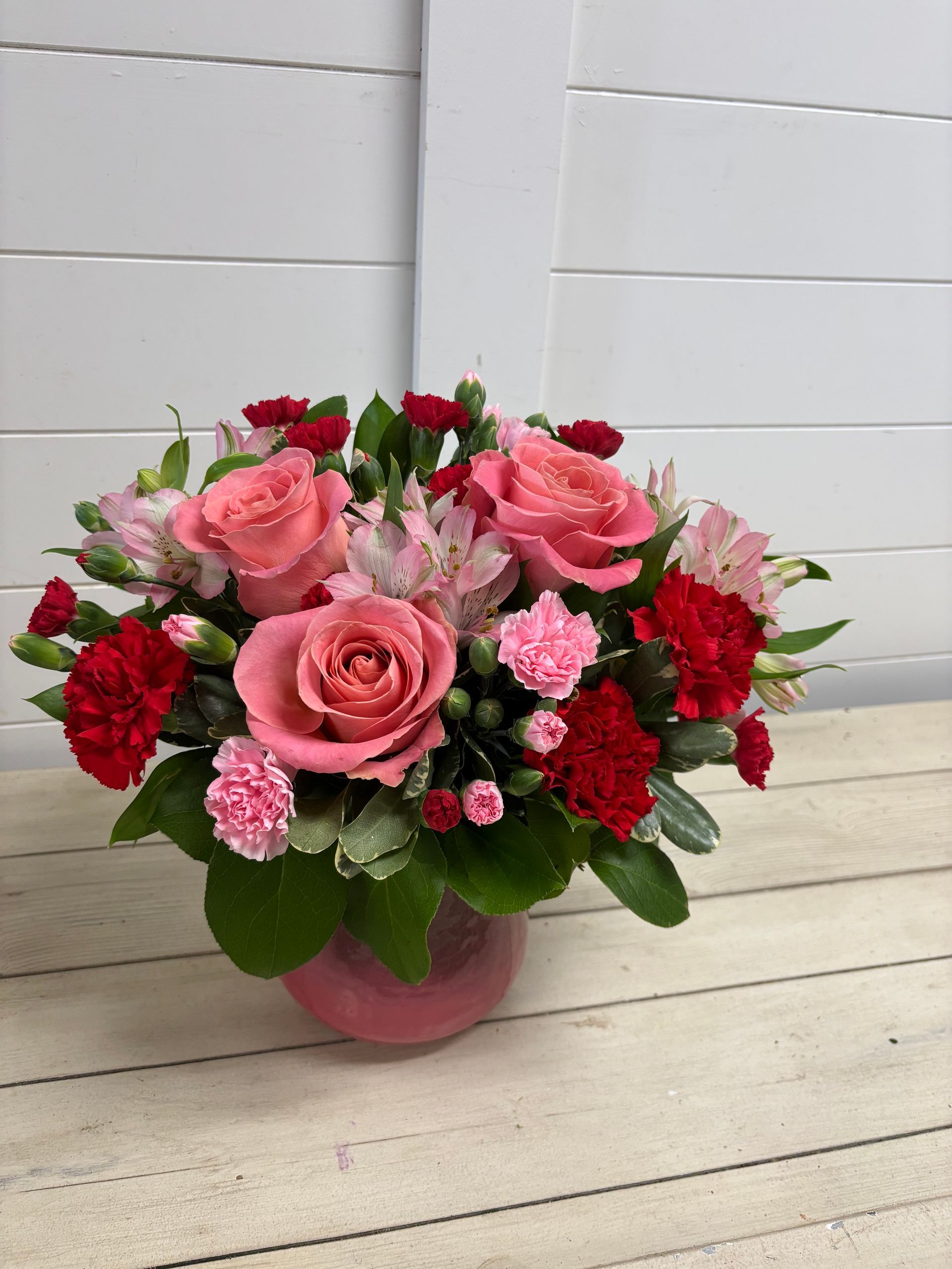 A floral arrangement with pink roses, red carnations, and light pink alstroemeria in a pink glass vase on a wooden surface.