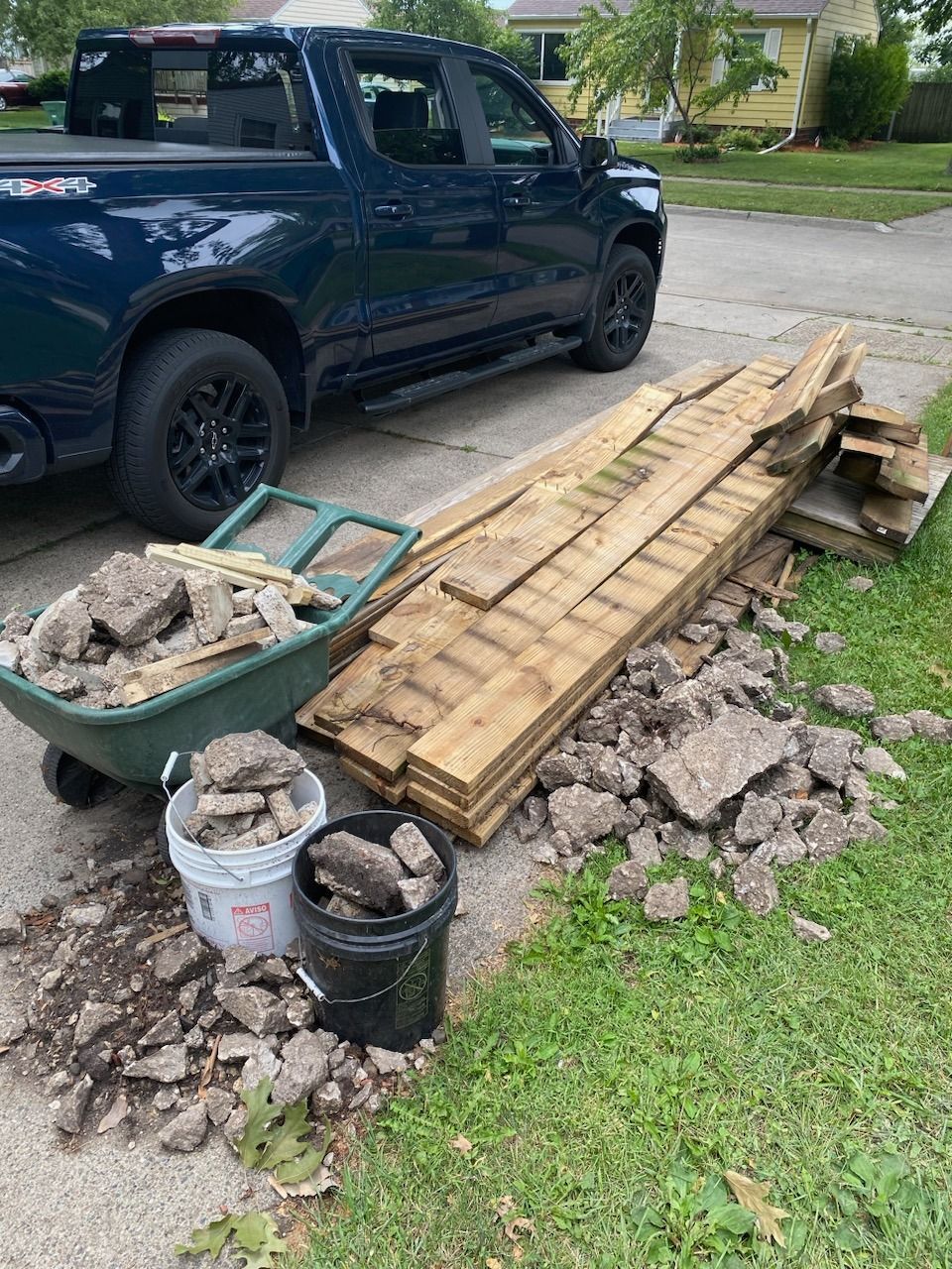 A blue truck is parked next to a wheelbarrow filled with rocks and wood.