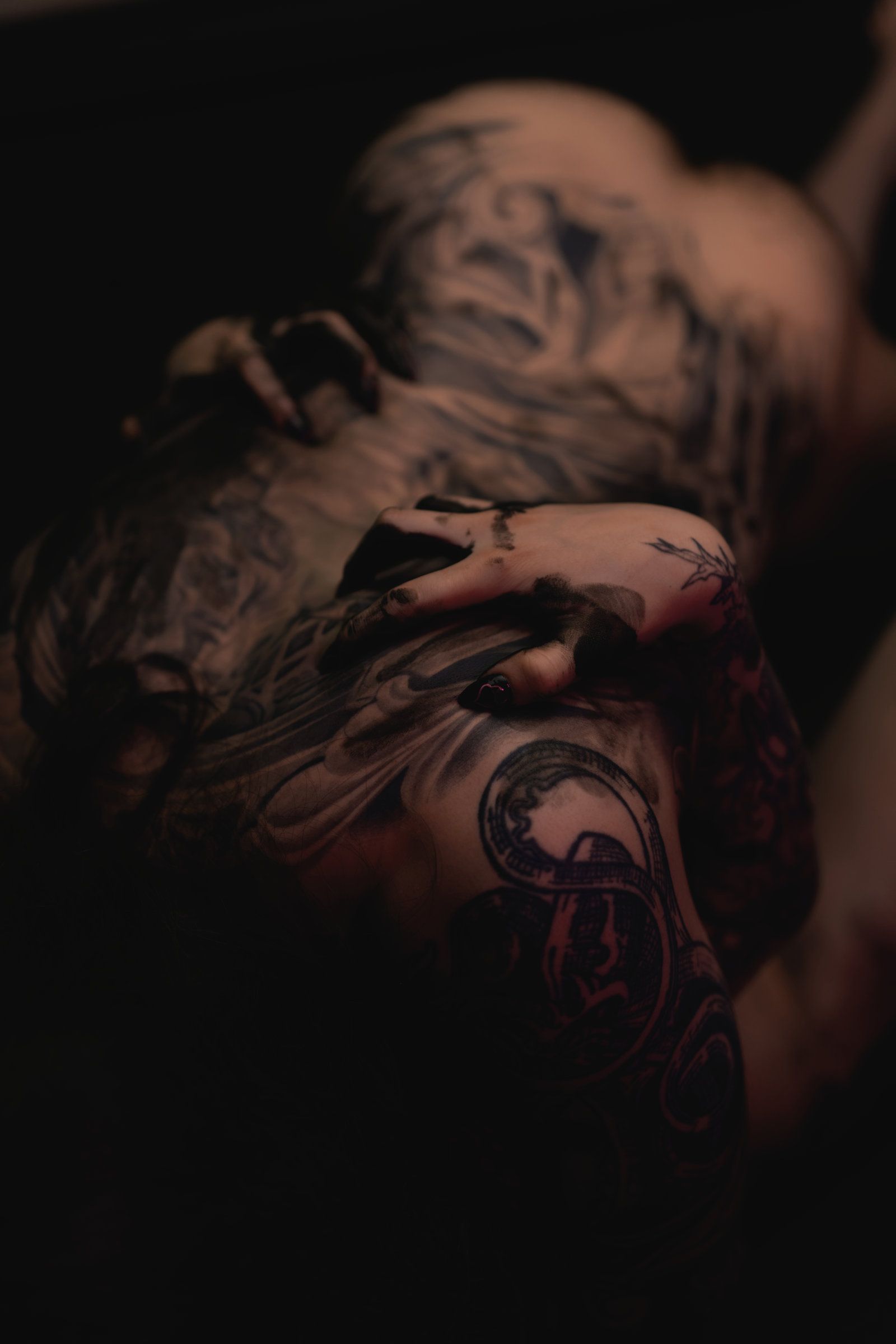 Personne avec des tatouages ​​complexes allongée, le visage caché, les mains près du visage, cadre sombre.