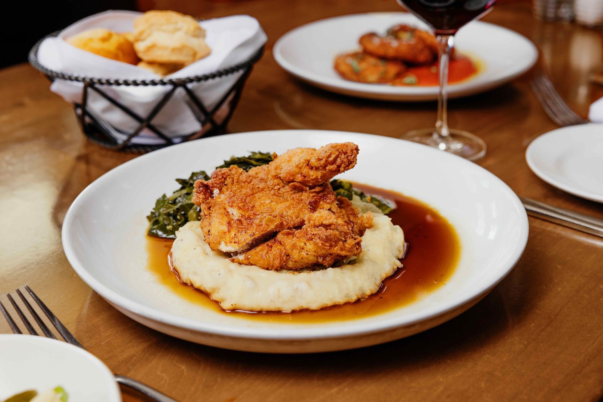 Fried chicken, mashed potatoes, and greens on a white plate with biscuits and wine at a restaurant.