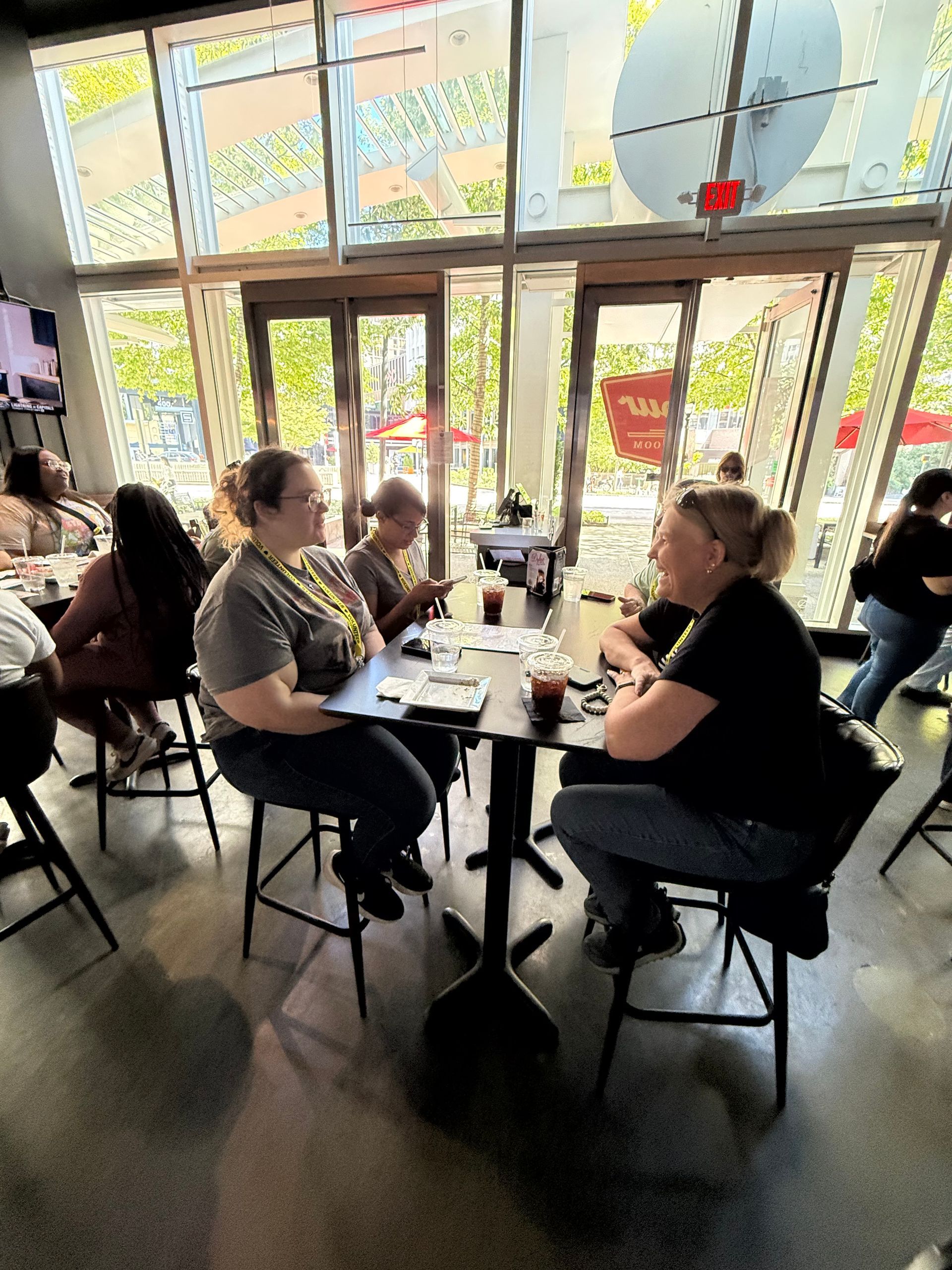 Two people seated at a table, conversing in a restaurant setting with others nearby. Large windows and doors are visible.