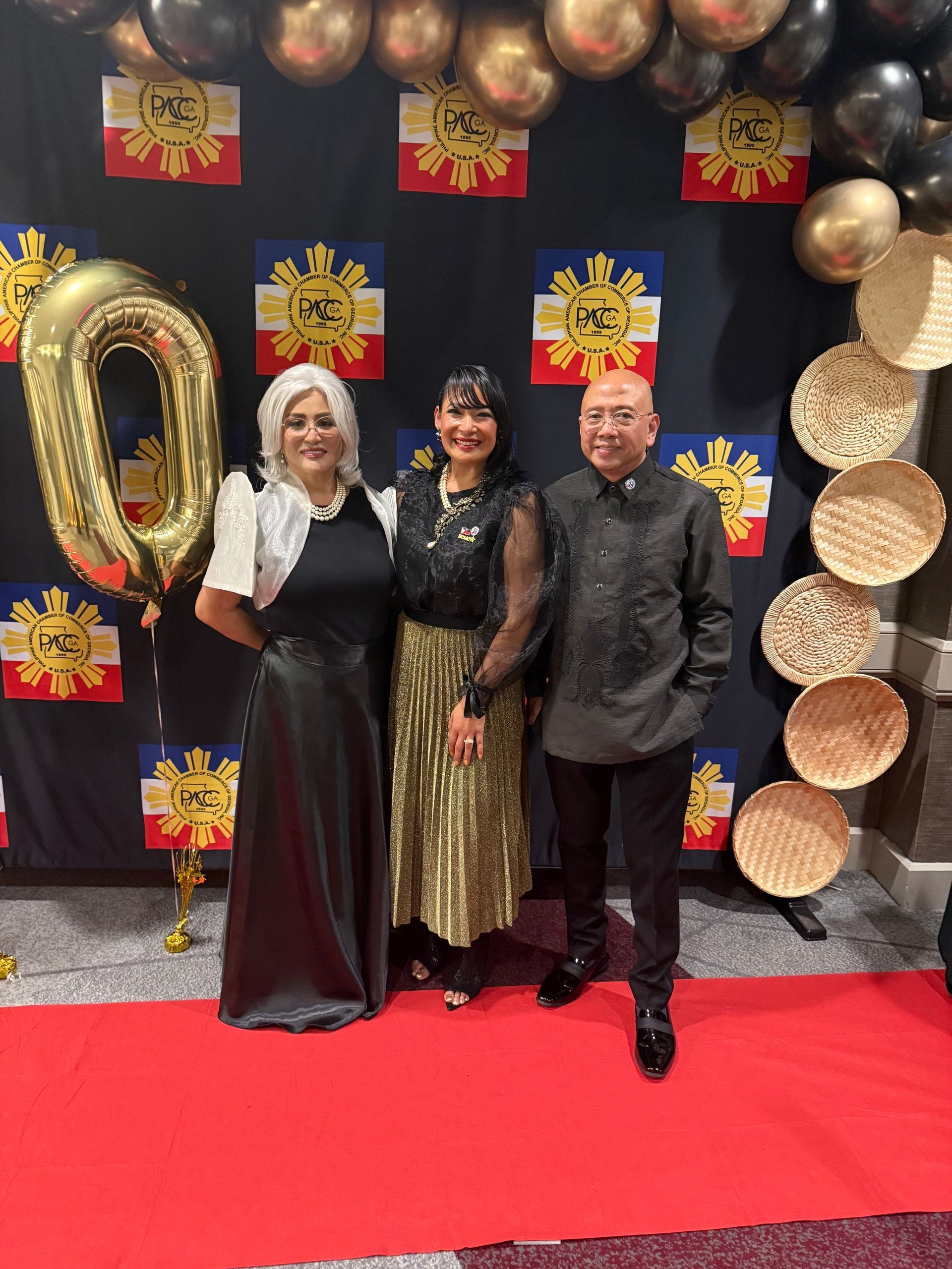 Three people in formal attire pose in front of Philippine flag decor at a party.