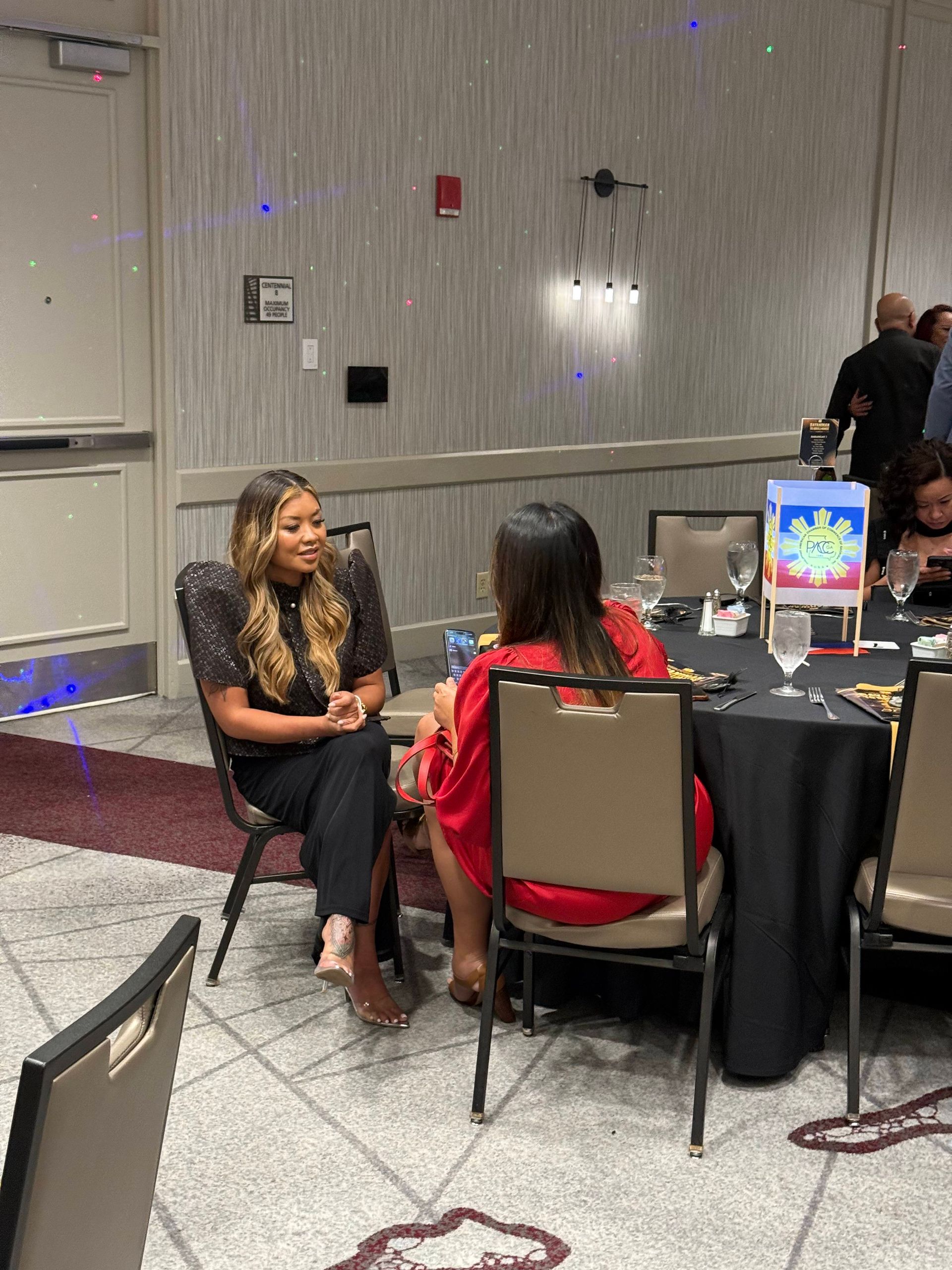 Two women seated at a table, conversing at an event. Dark attire, black tablecloth, festive decorations.