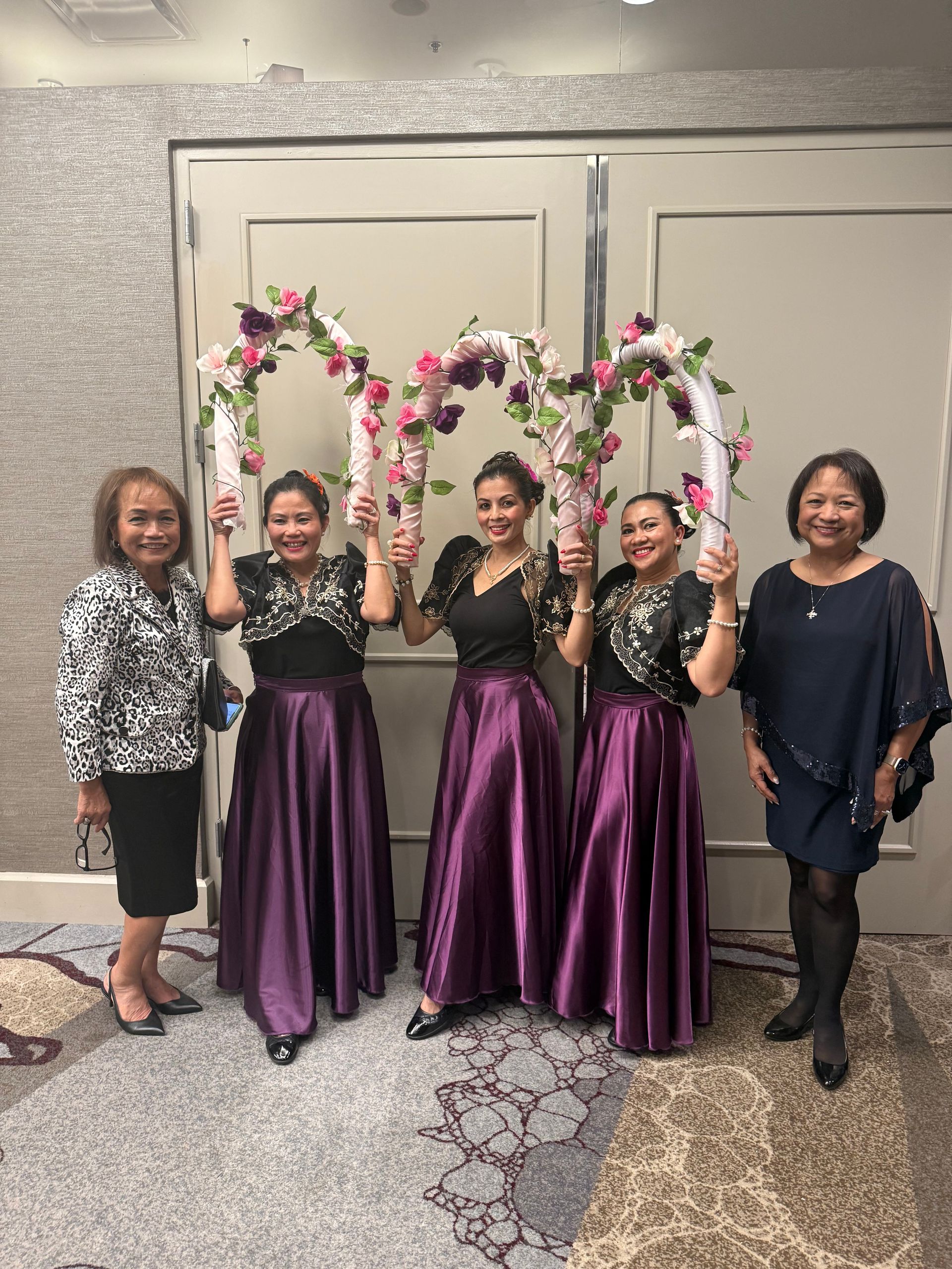 Five women pose with floral arches, wearing formal attire. Hotel interior.