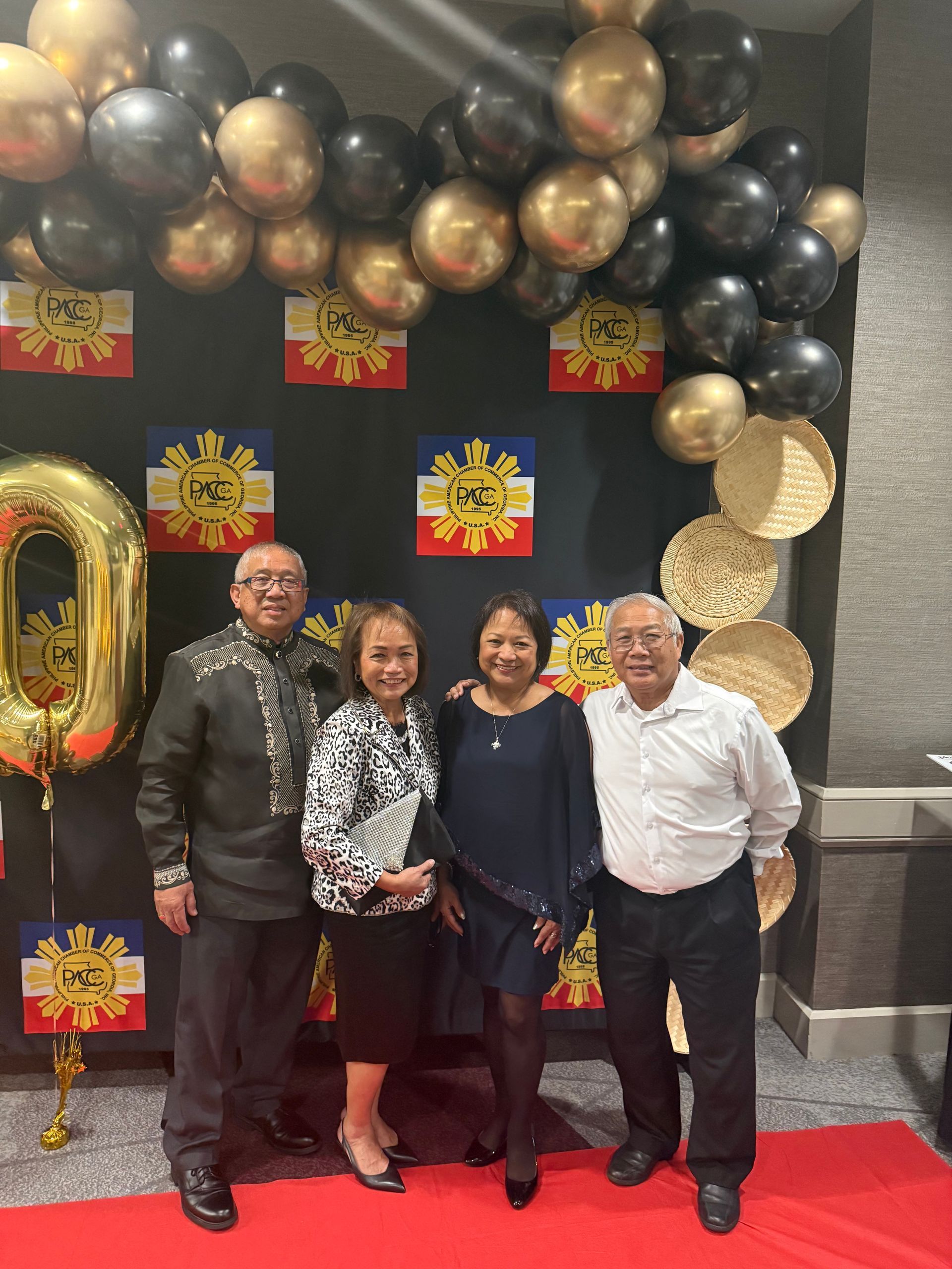 Four people pose for photo at a decorated event with gold and black balloons, Filipino flags, and a red carpet.