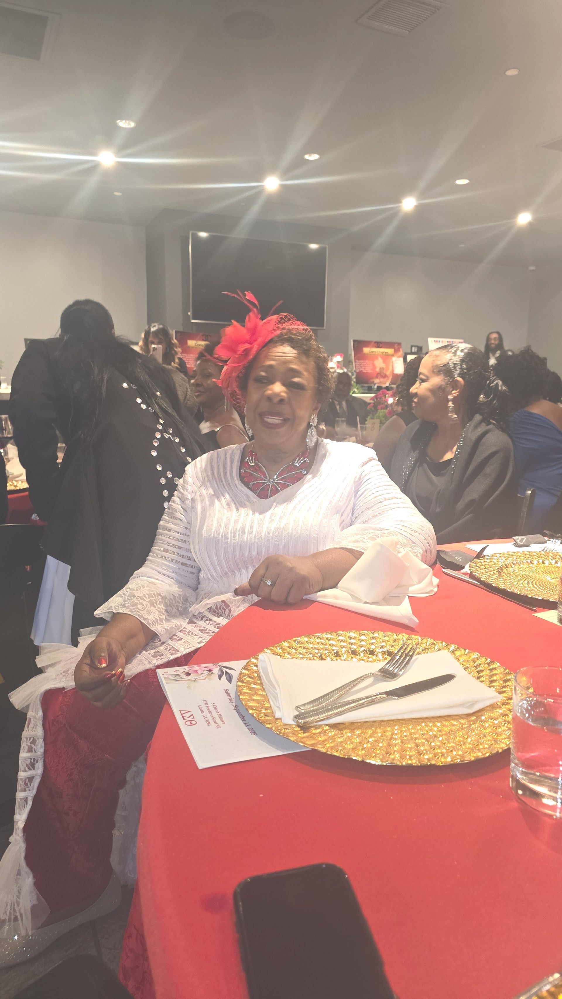 Woman seated at a red-clothed table, wearing a white lacy top and red floral headpiece; at an event.