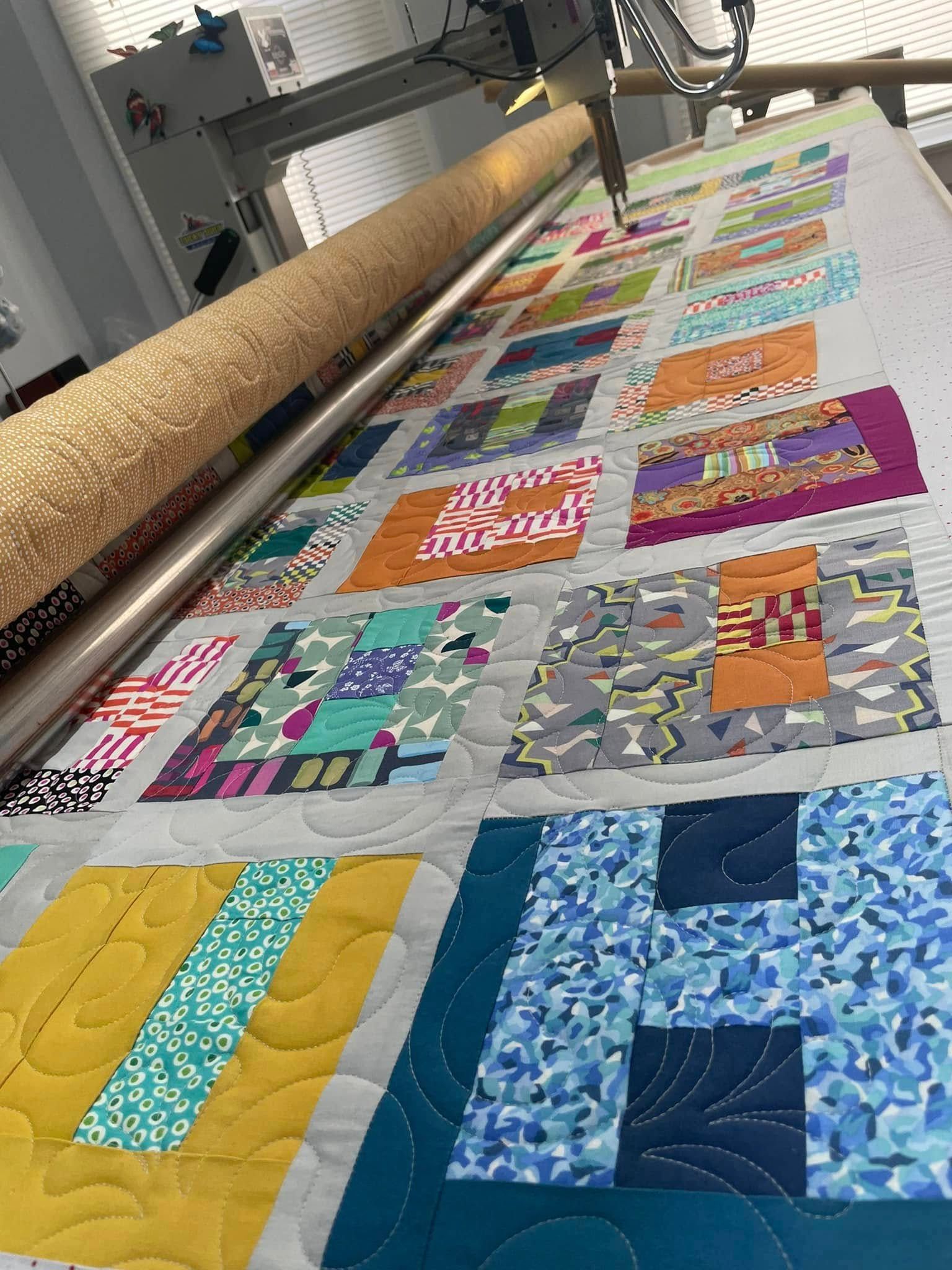 A long-arm quilting machine stitches a colorful, patchwork quilt top composed of various patterned fabric squares.