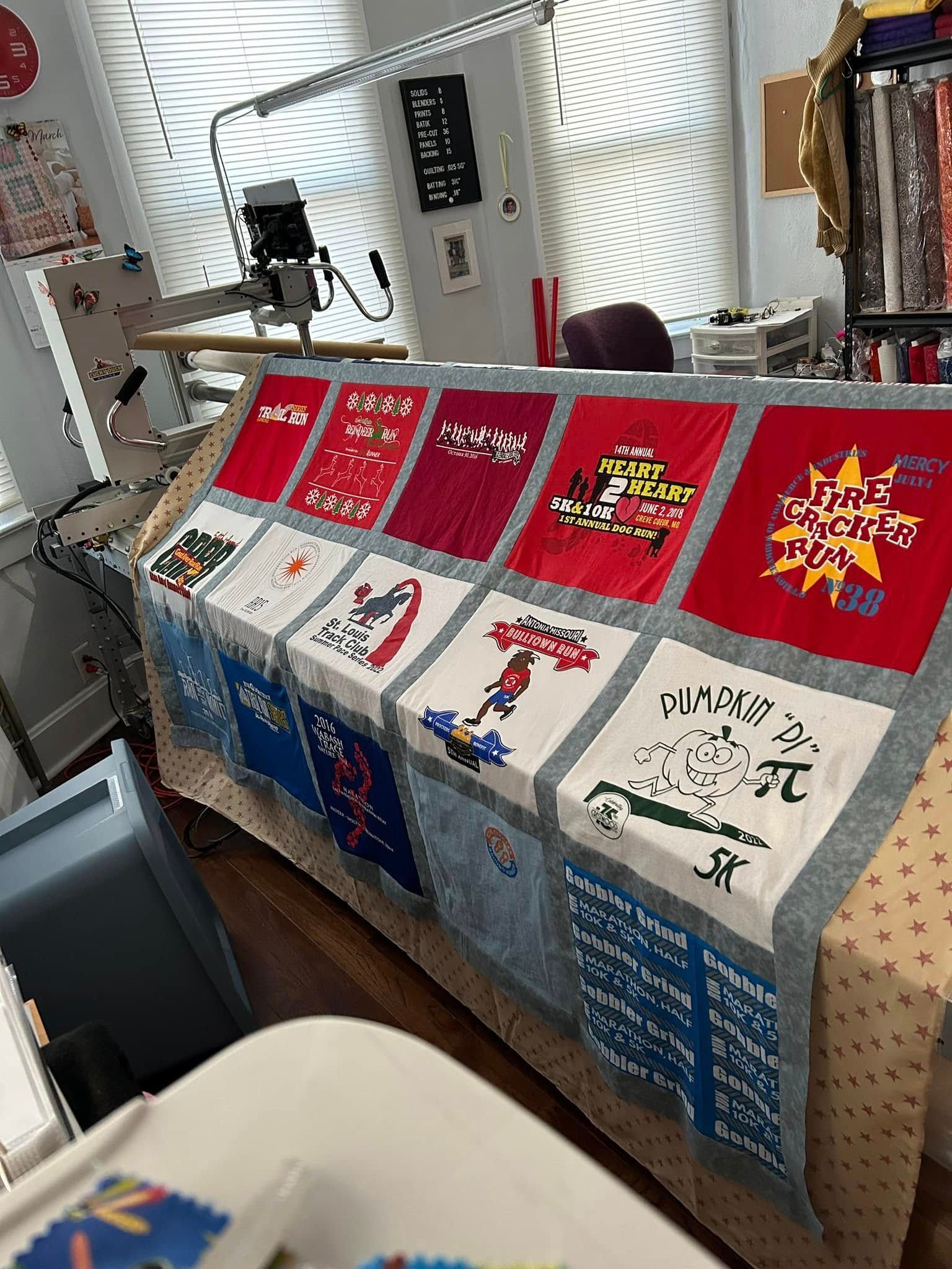 A t-shirt quilt being quilted on a longarm machine in a craft room with windows and fabric storage in the background.