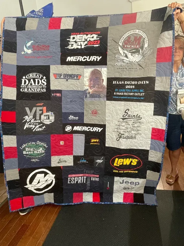 A person holds up a homemade t-shirt quilt made of various black, grey, and red fabric panels with different logos.