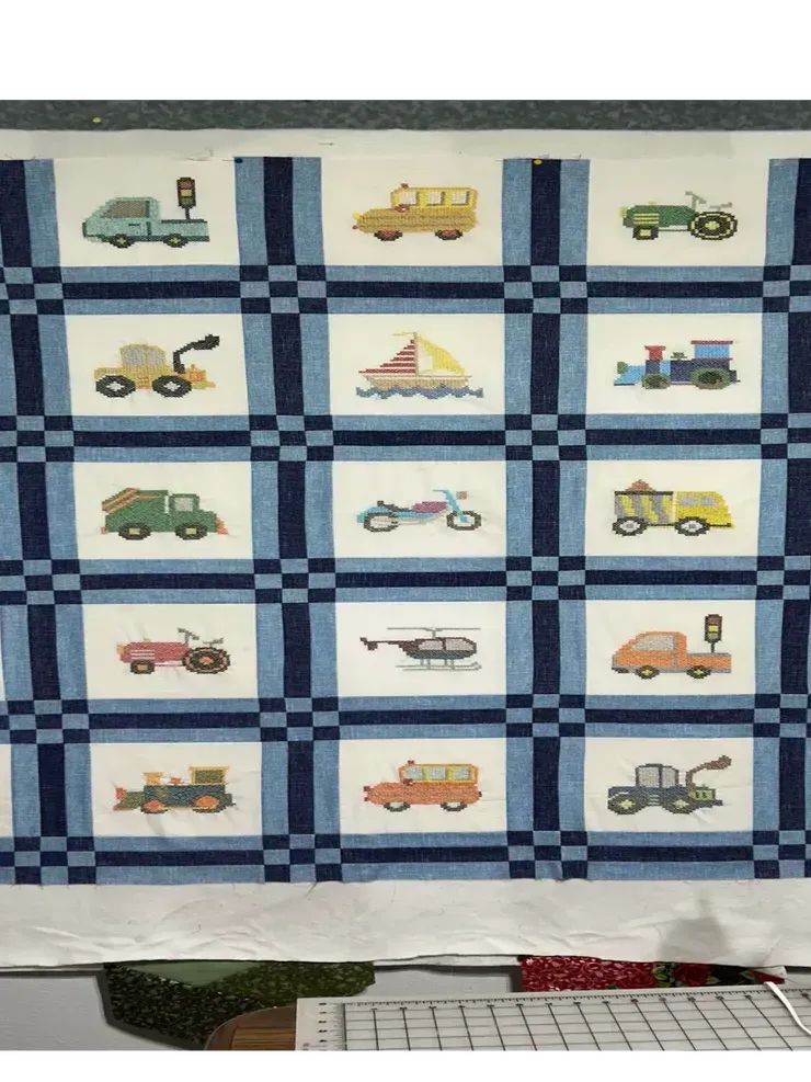 A quilt featuring a grid of fifteen blocks, each embroidered with a different colorful vehicle, set in blue sashings.