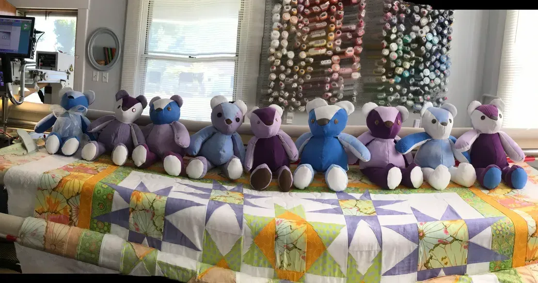 Nine handmade teddy bears in shades of blue and purple sit in a row on a quilted blanket in a craft room.