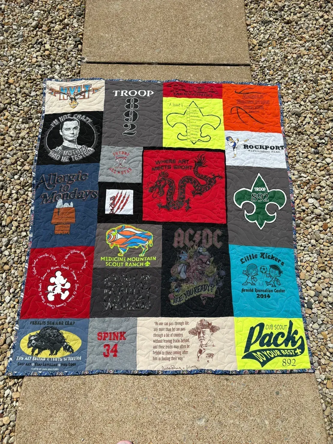 A T-shirt quilt featuring various graphic panels, including scouting logos, band shirts, and Disney designs, on a path.