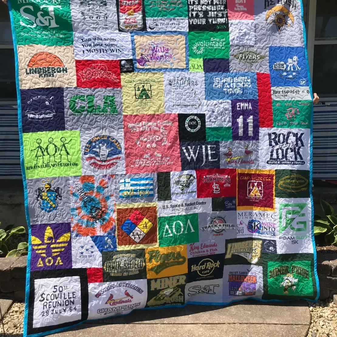 A colorful t-shirt quilt composed of numerous square patches with various logos, text, and graphics, bordered in bright blue.