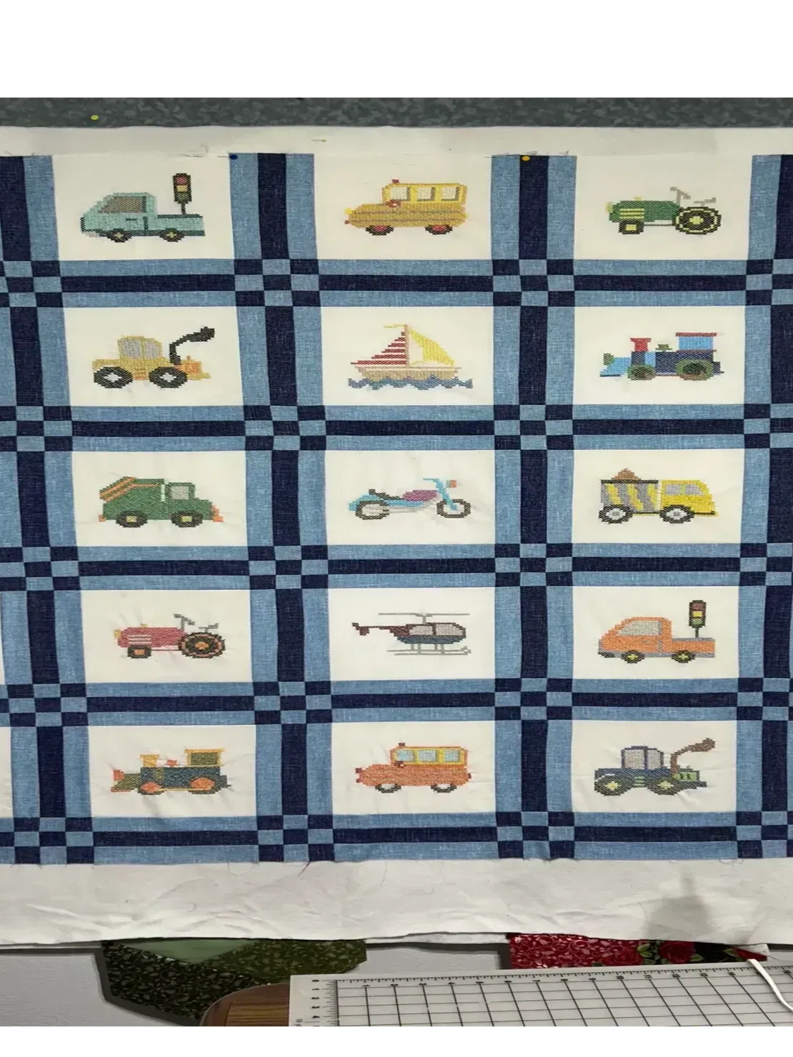 A quilt featuring a grid of fifteen white squares, each depicting a different embroidered vehicle, separated by blue borders.