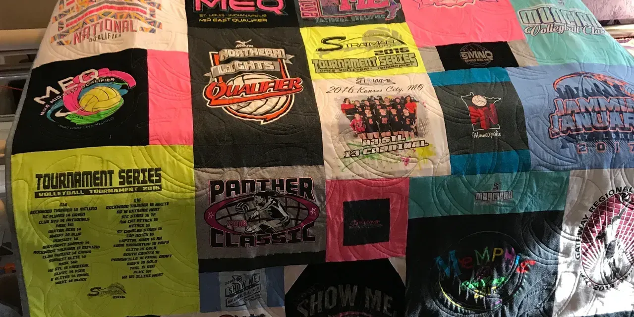 A colorful quilt made from various sports and event T-shirts, arranged in a grid-like pattern on a bed.