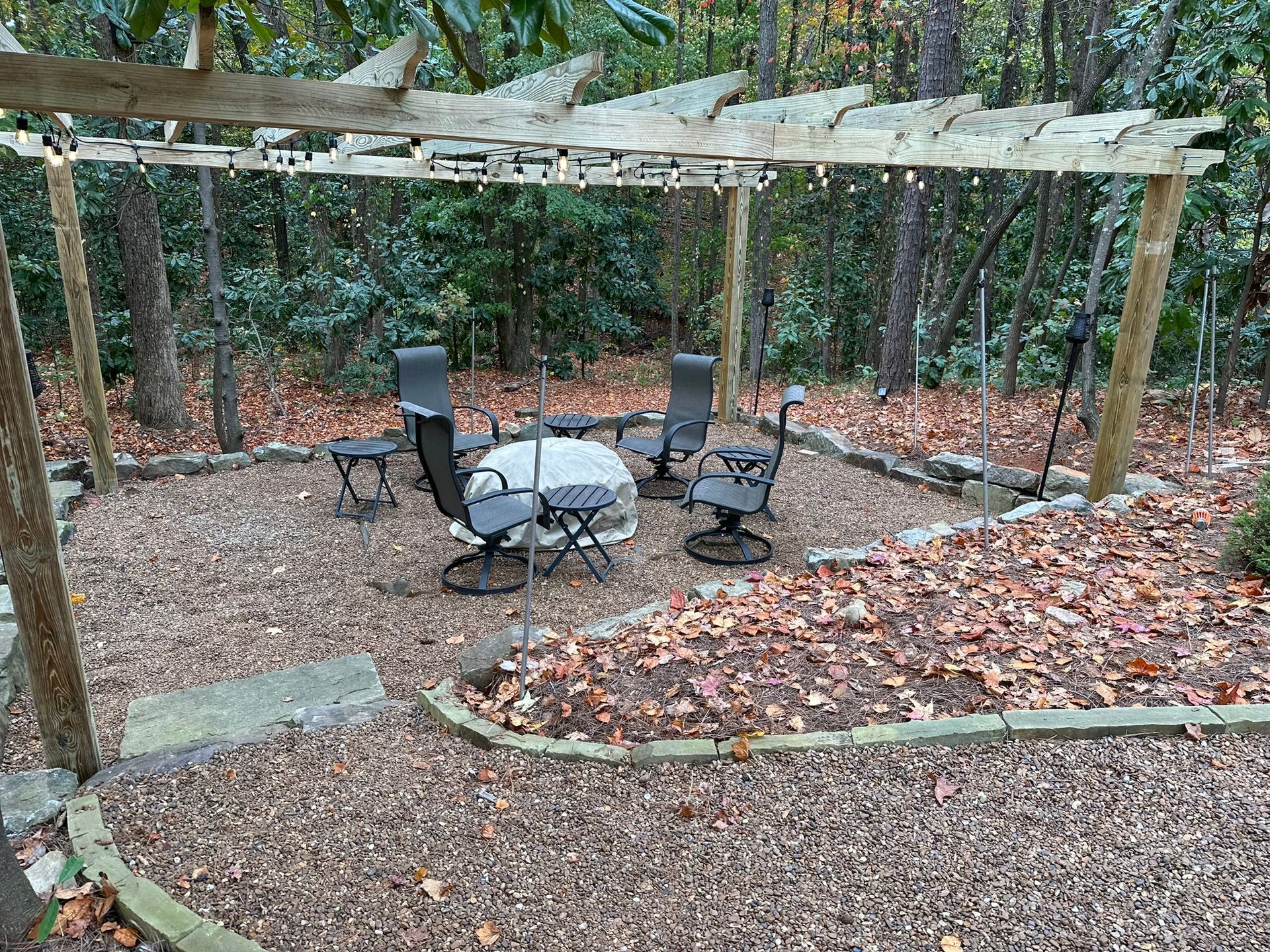 A patio with a fire pit and chairs under a pergola in the woods.
