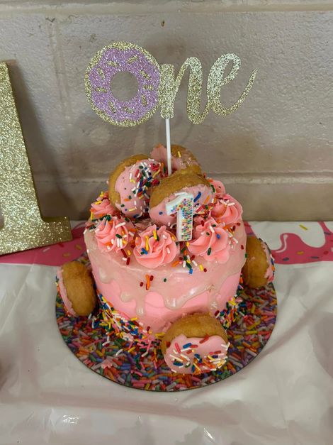 Pink drip cake with sprinkles and a glittery “one” topper on a decorated cake board