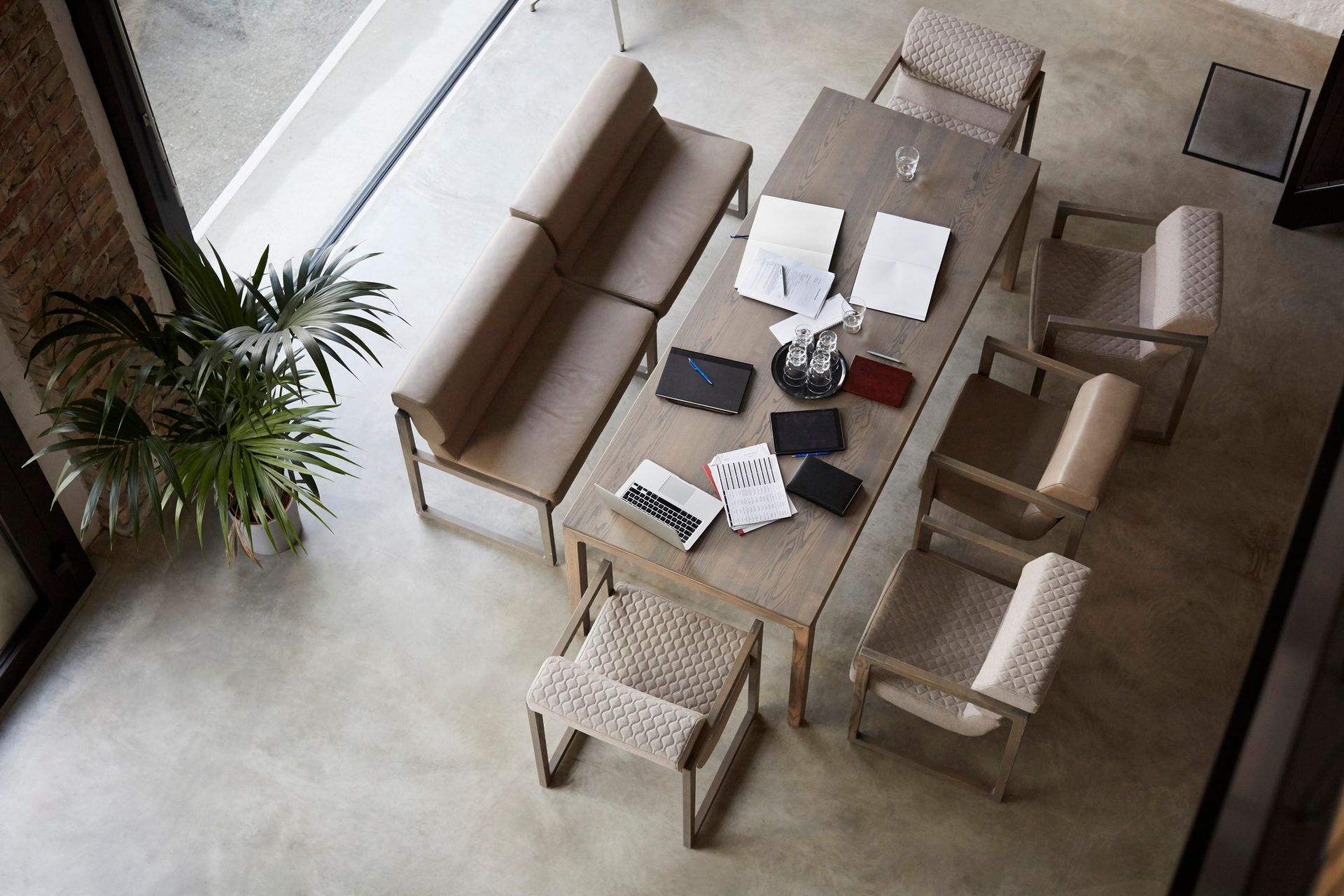 High-angle shot of a modern dining room with a long wooden table, chairs, and a bench. There are papers, notebooks, and a small plant on the table.