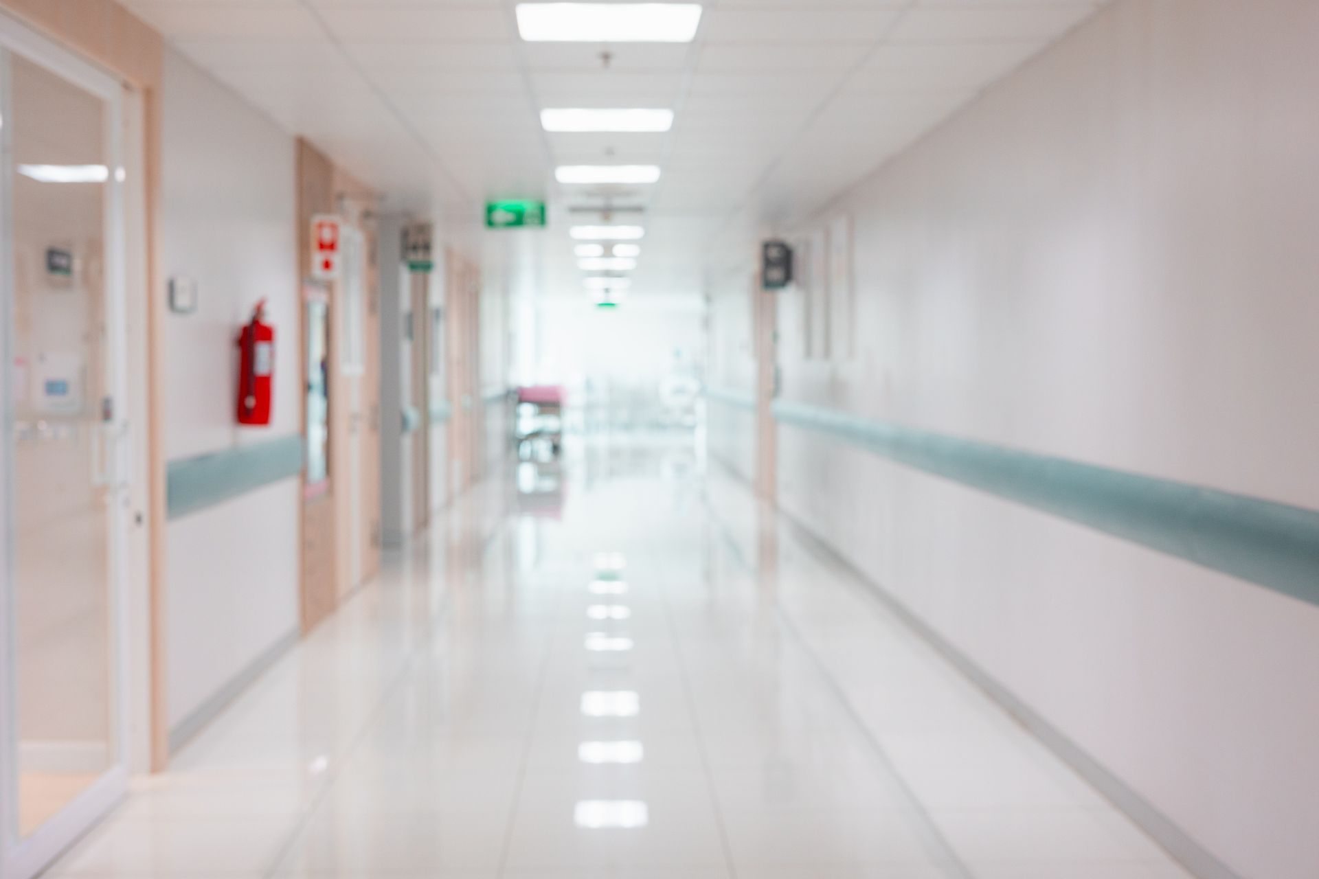 Blurred view of a brightly lit hospital hallway with white walls, a polished floor, and a gurney in the distance.