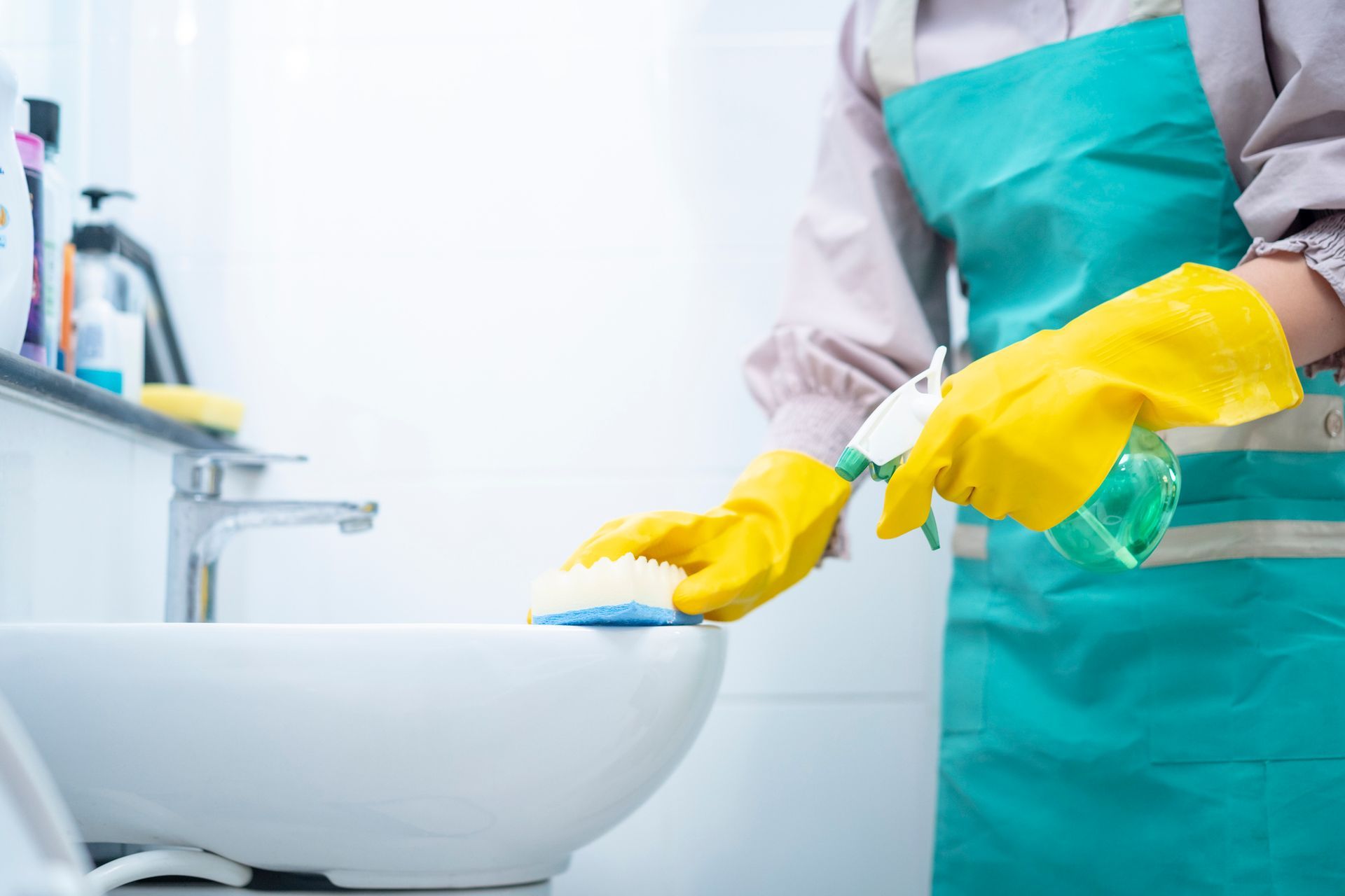 Person wearing yellow gloves and a green apron cleans a white sink with a sponge and spray bottle in a bathroom.