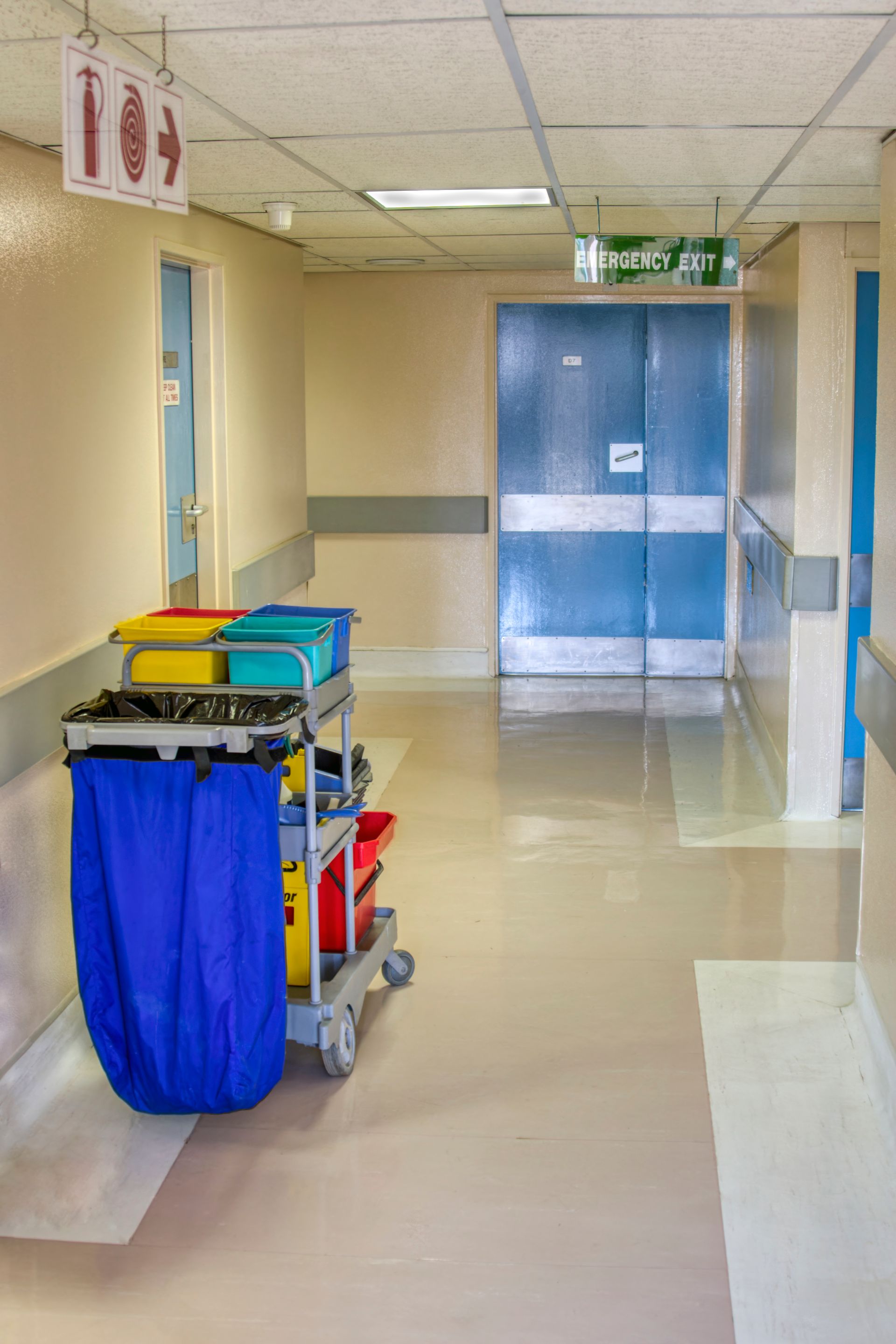 A hospital hallway with a cleaning cart, emergency exit sign, and a blue door.