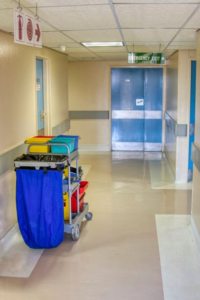 A hospital hallway with a cleaning cart, emergency exit sign, and a blue door.
