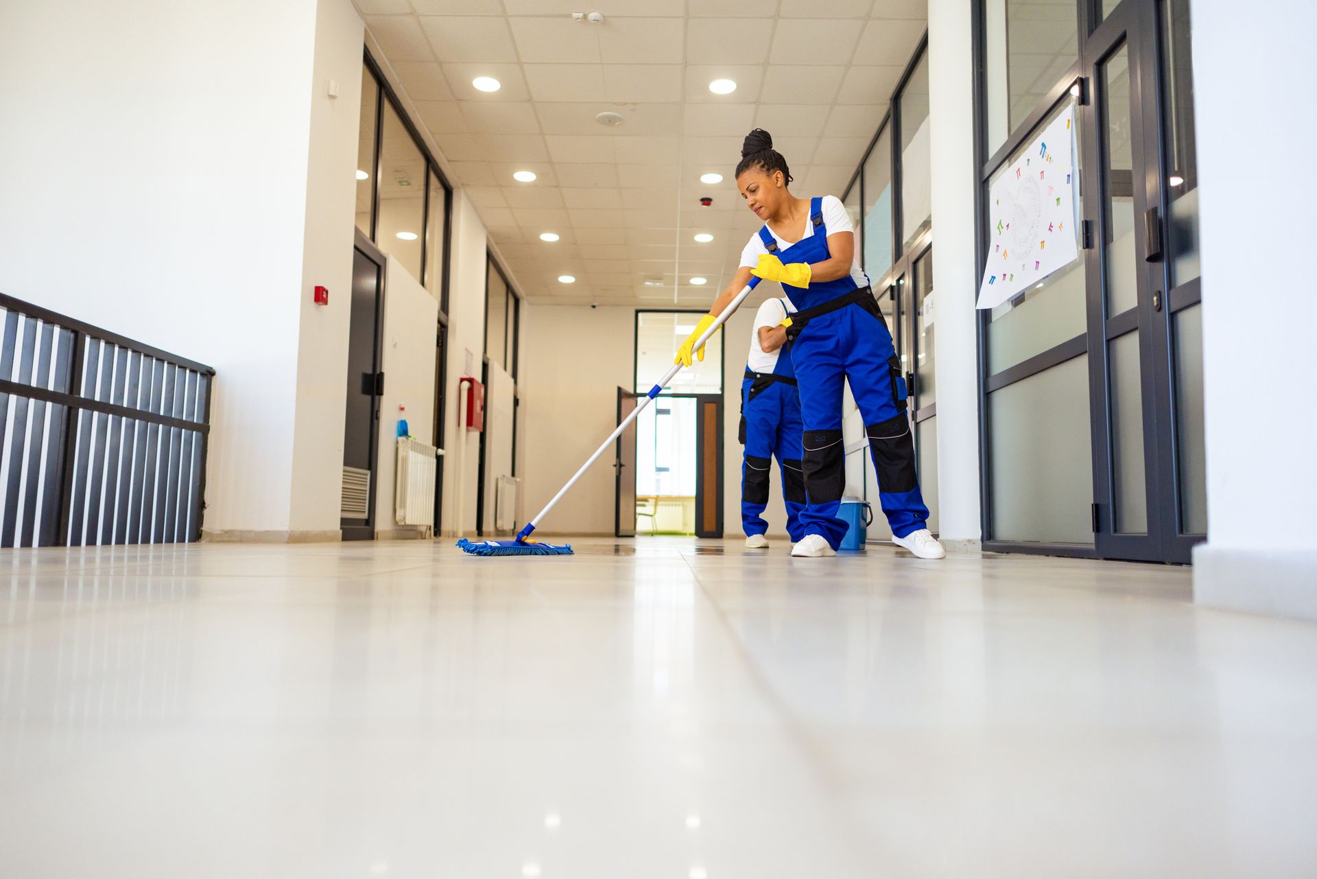 Two people in blue work uniforms clean a bright, long hallway. One mops the floor while the other stands nearby.