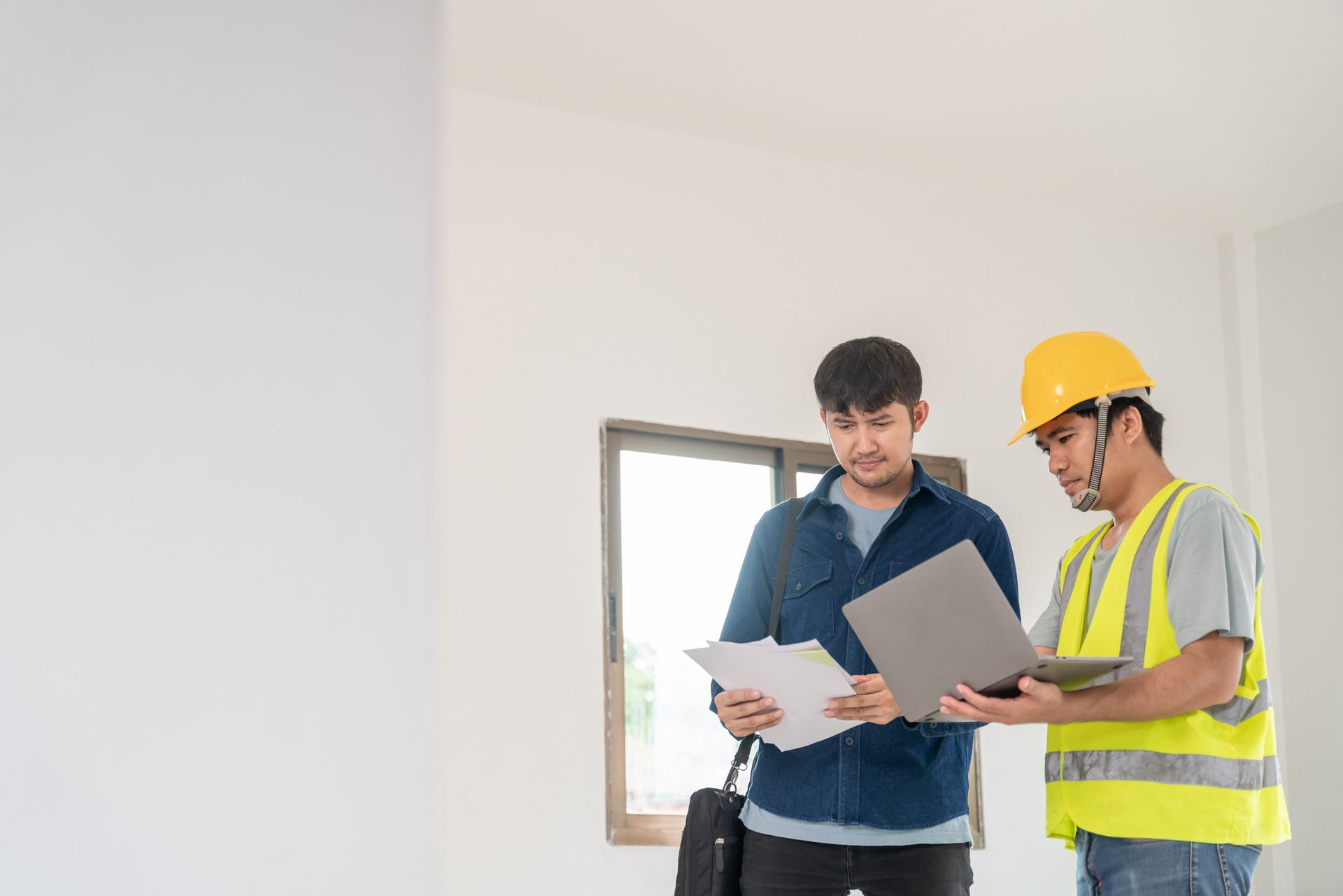 Two men reviewing documents and a laptop inside a room. One wears a hard hat and safety vest.