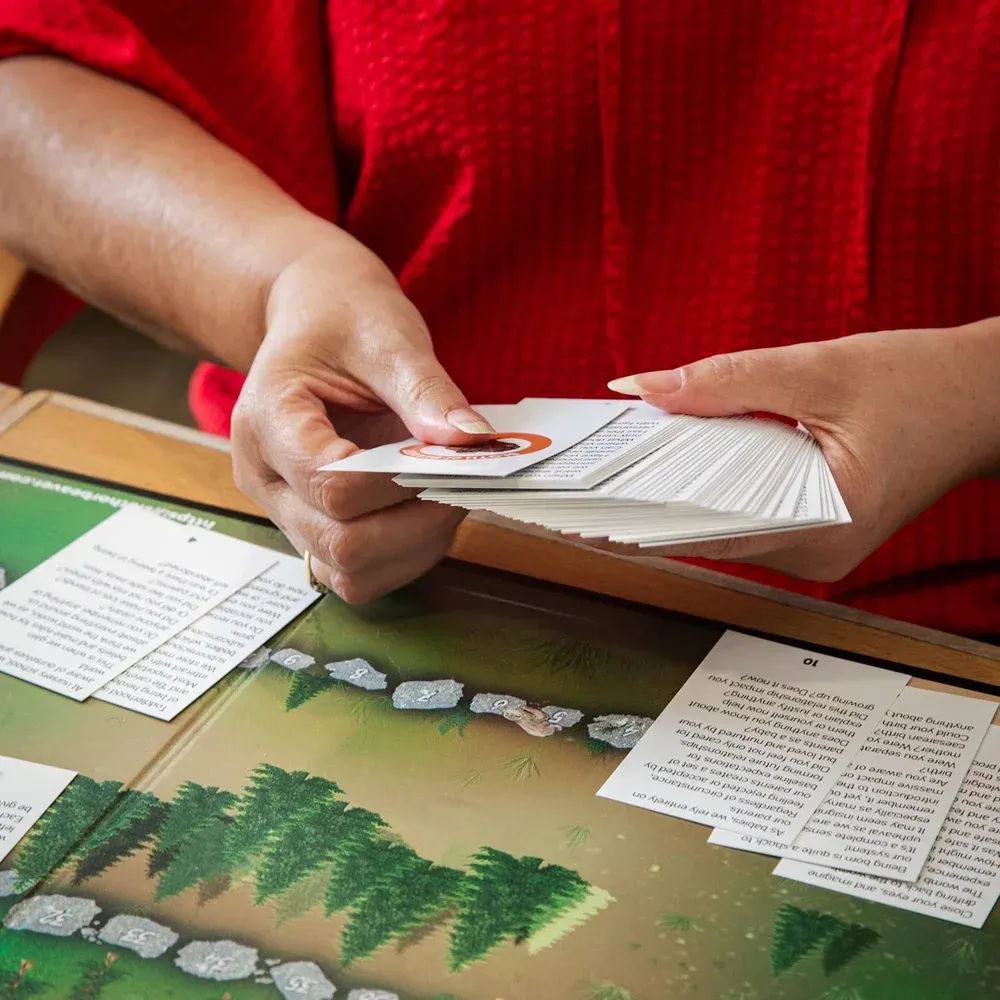 A person in a red shirt holds a deck of cards over a forest-themed board game with playing cards spread on the surface.