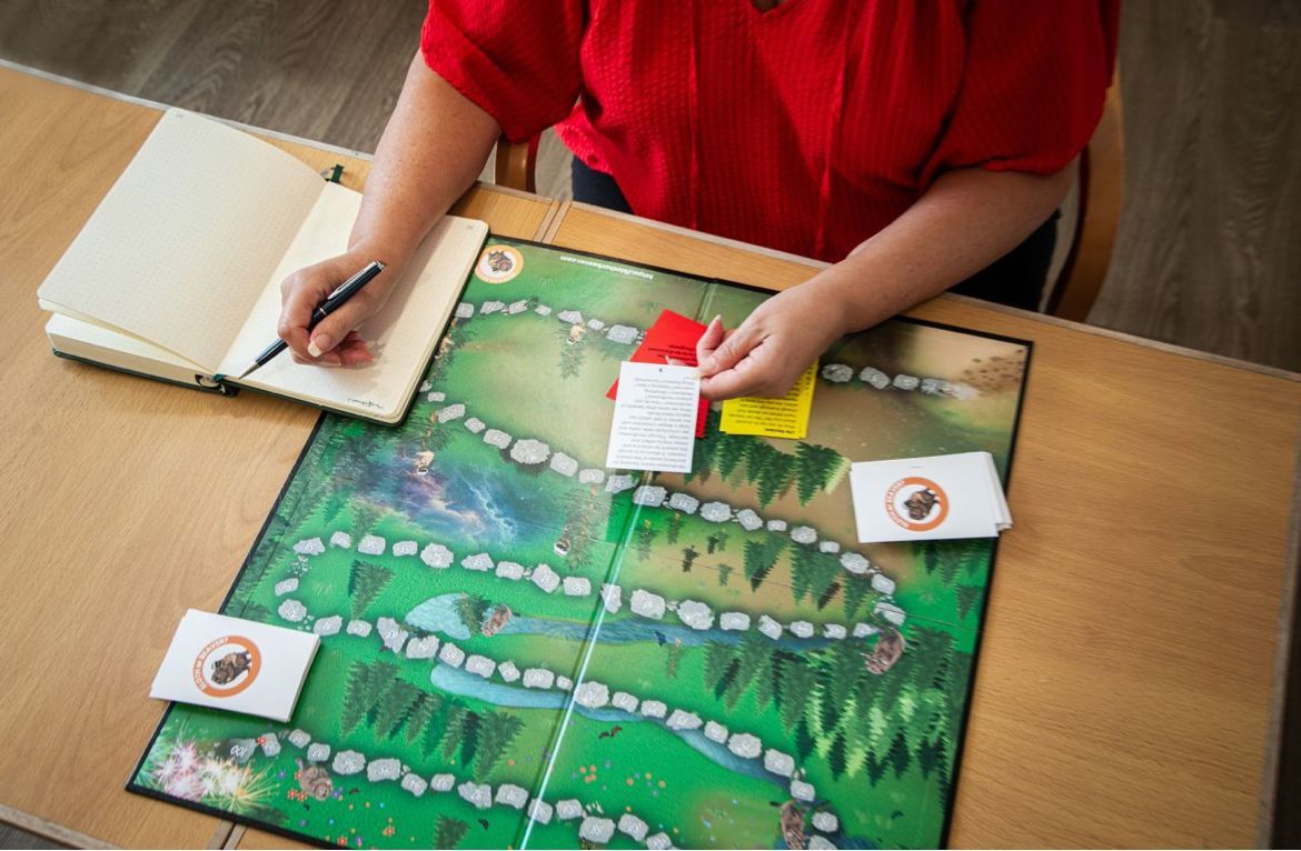 A person in a red shirt takes notes while playing a board game with stone paths, cards, and markers on a wooden table.