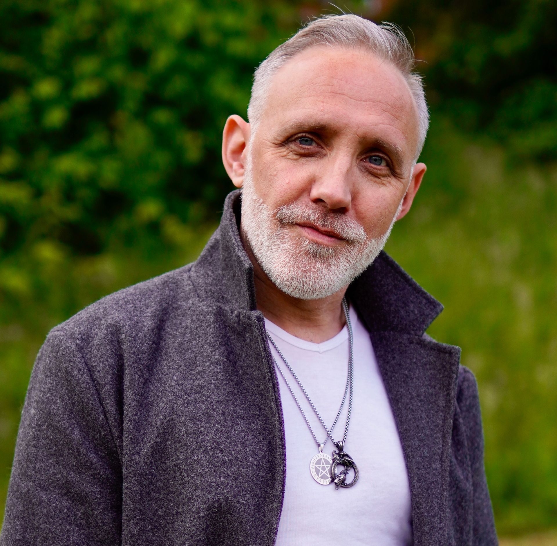 A man with a salt-and-pepper beard wearing a gray coat and two necklaces against a blurred green outdoor background.