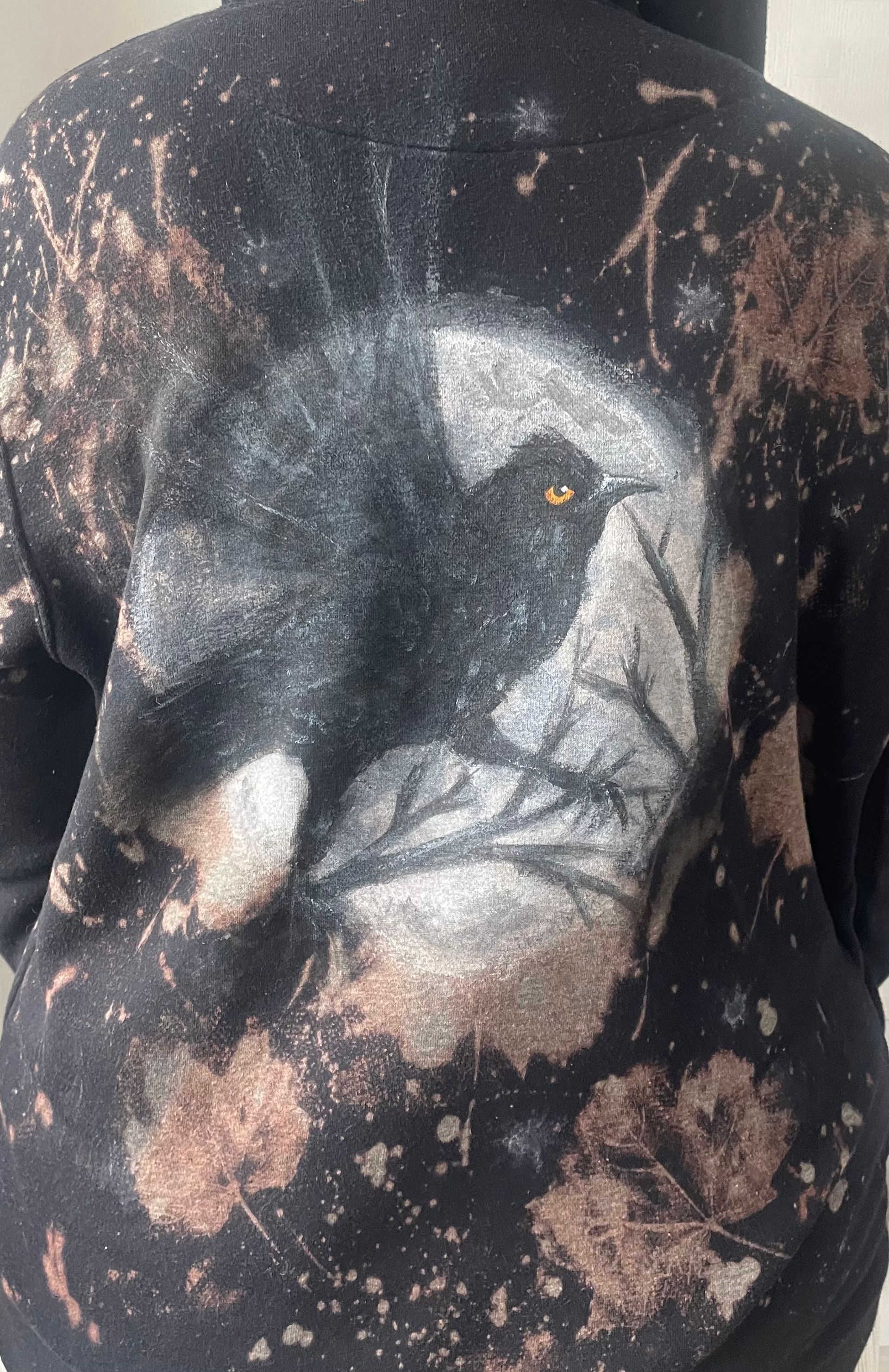A black hooded sweatshirt with a painting of a crow against the full moon on the back, with bleached leaf prints all around
