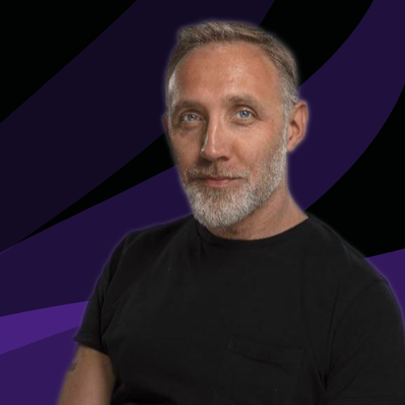 A man with a beard is wearing a black shirt and sitting in front of a purple background.