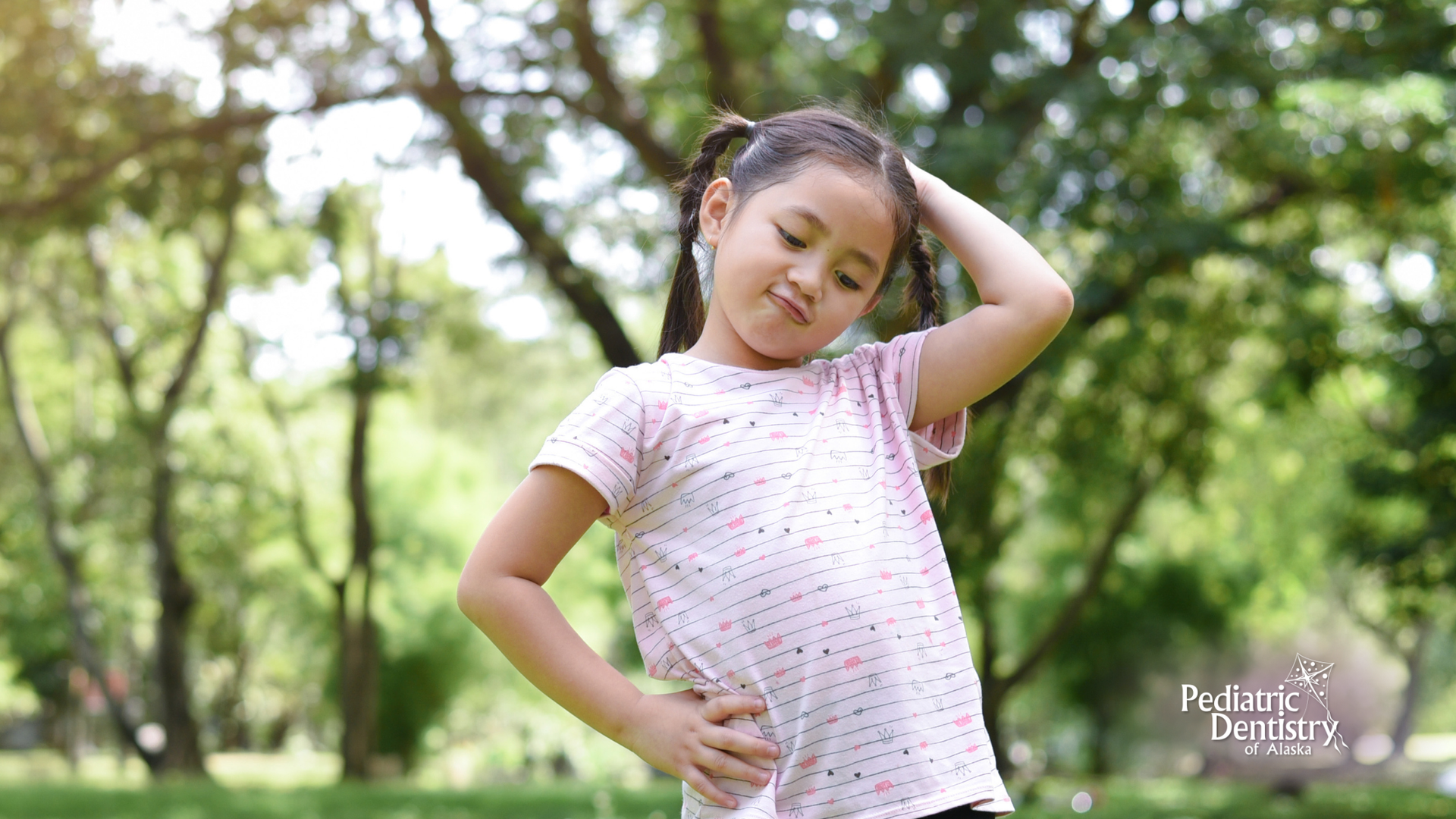 A little girl is standing in a park with her hands on her hips.