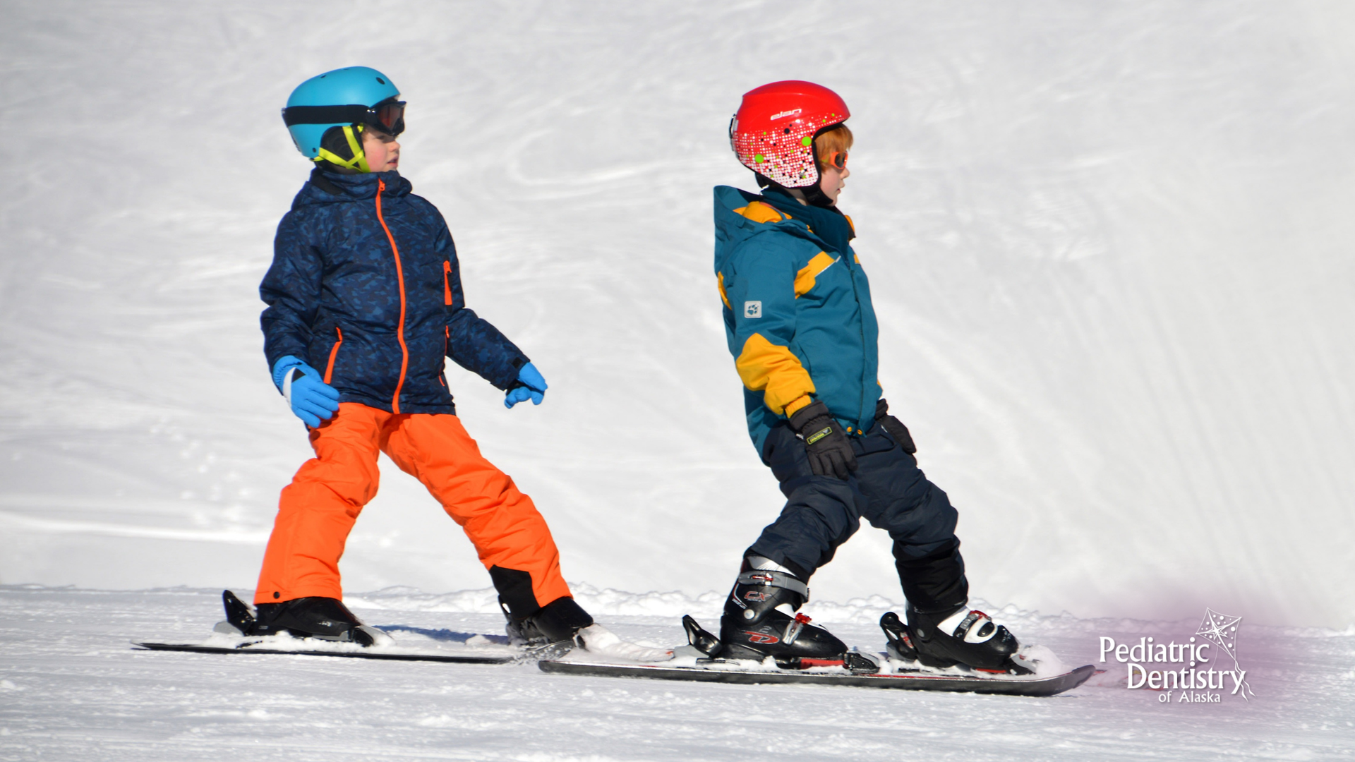 Two young boys are riding snowboards down a snow covered slope.